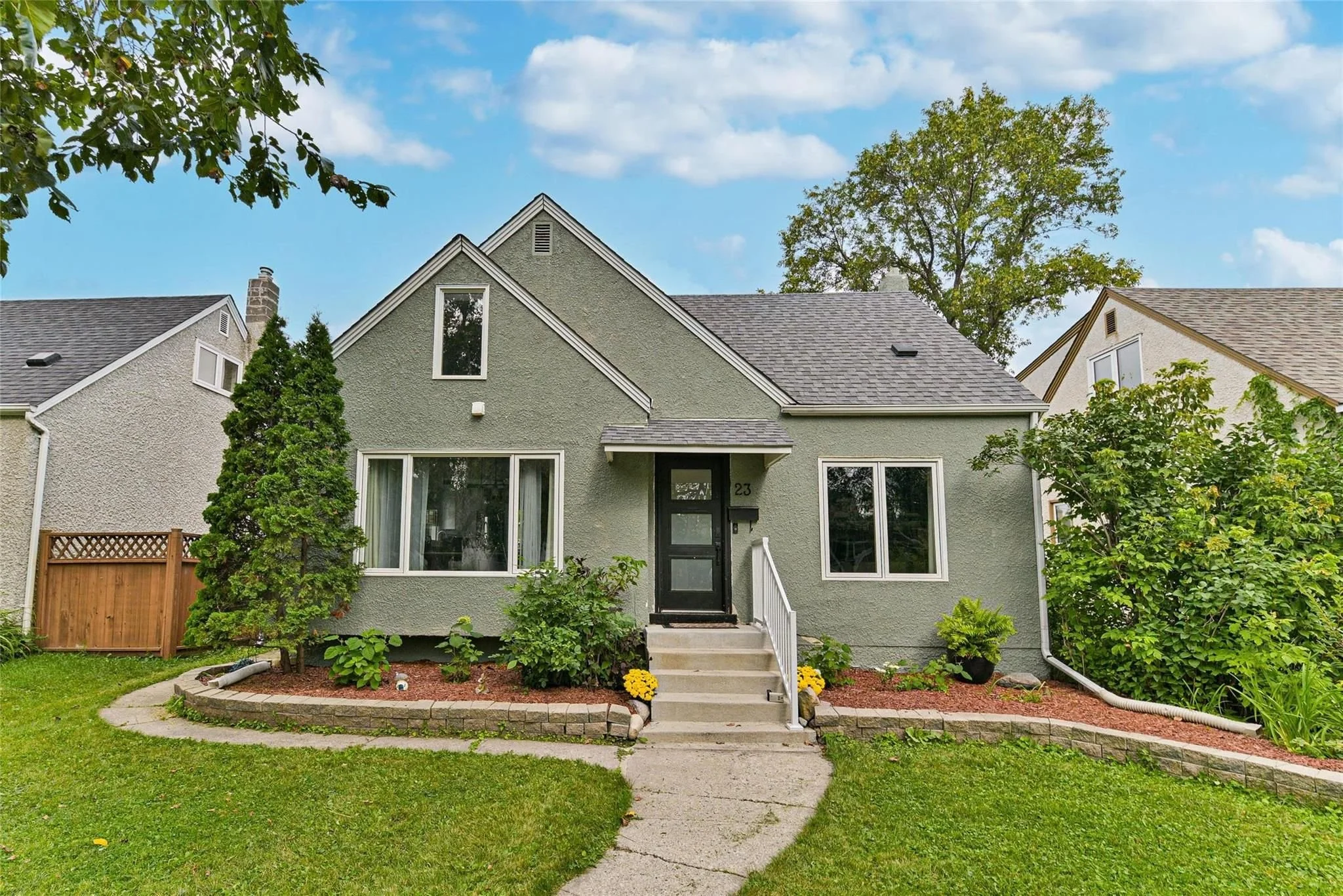 A light green house with white trim and a black front door, surrounded by a lawn and some plants, with neighboring houses on both sides, under a partly cloudy sky.