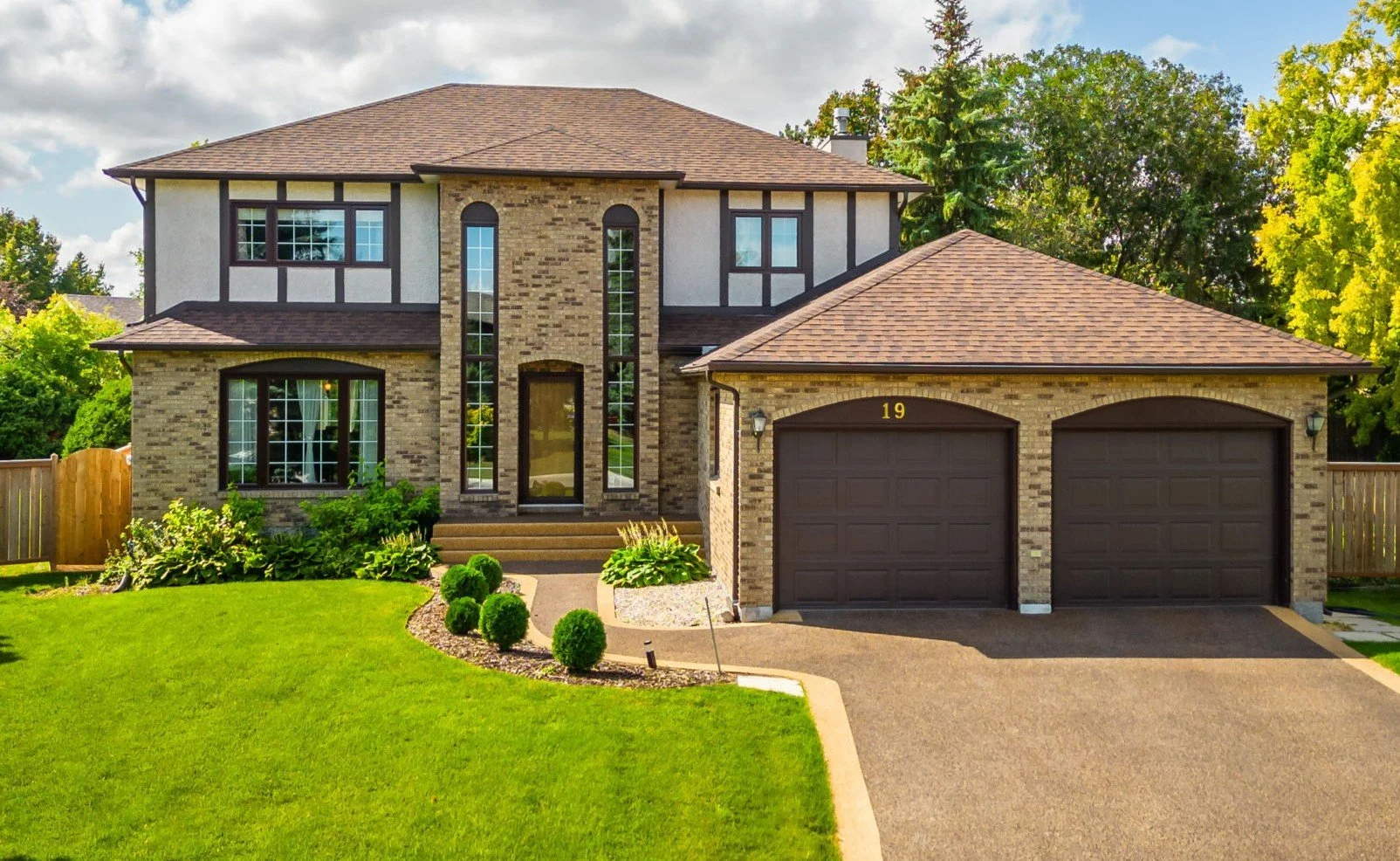 A two-story house with brick and siding exterior, brown roof, large front windows, and a double garage. The front yard has a green lawn, small bushes, and a walkway leading to the front door. The house number is 19.