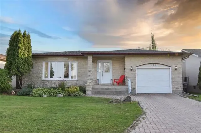 A single-story house with a brick exterior, white front door, and attached garage. The house has a lawn with a tree on the left and a paved driveway. There are two red chairs on the front steps and a sunset sky in the background.