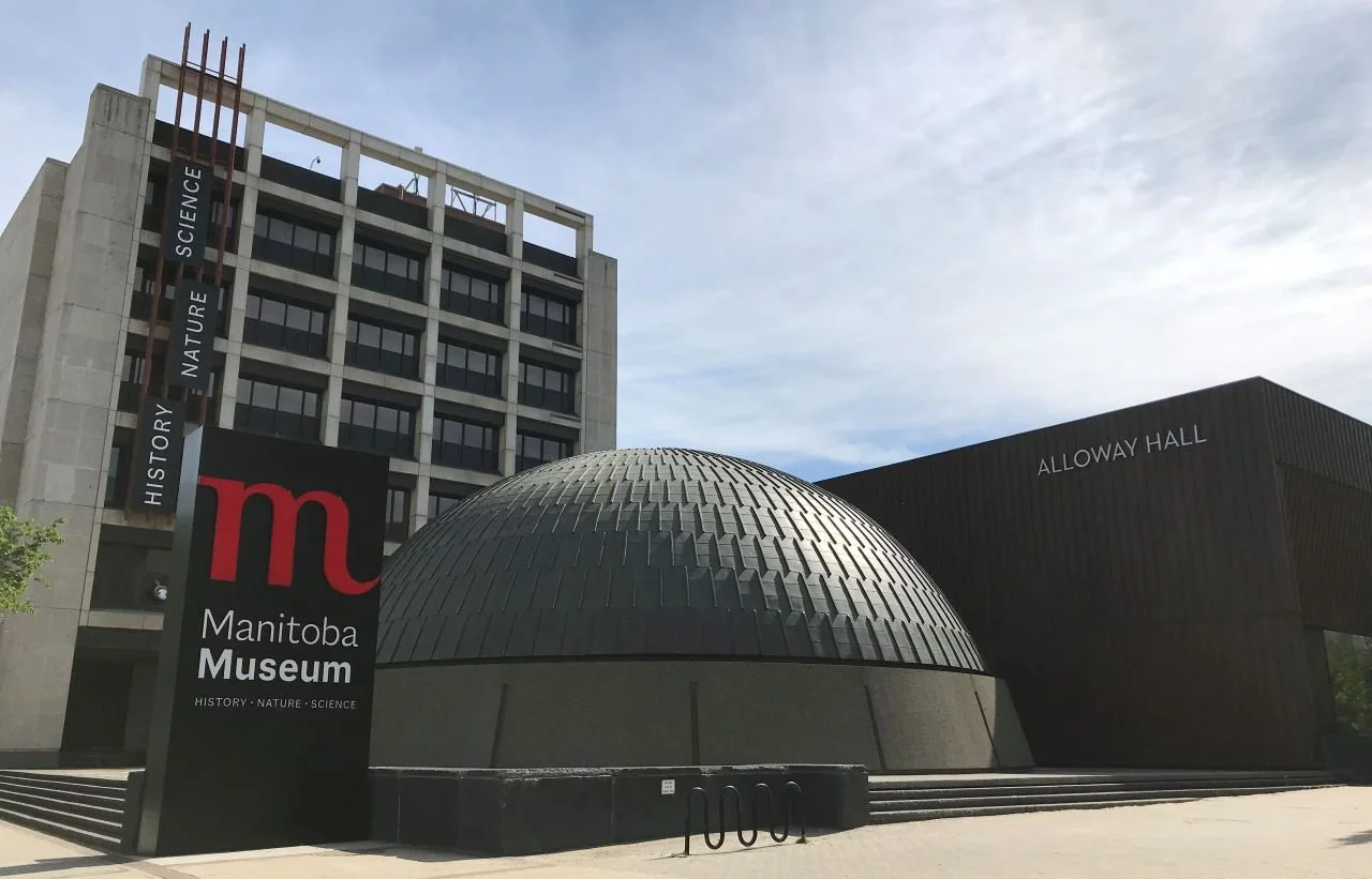 Exterior view of the Manitoba Museum with a sign that reads 'Manitoba Museum' and shows the categories 'History,' 'Nature,' and 'Science.' The building features a large, rounded, dome-like structure and a rectangular section labeled 'Alloway Hall,' with a modern multi-story building in the background. The sky is partly cloudy.