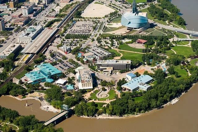 Aerial view of an urban park with a large modern building with a domed roof, surrounding green areas, pathways, and a river at the bottom edge.