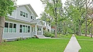 A white house with large windows, surrounded by green trees and a well-maintained lawn, with a sidewalk leading to the entrance.