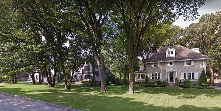 Quiet suburban neighborhood with large green trees and well-maintained lawns, featuring two houses with traditional architecture.