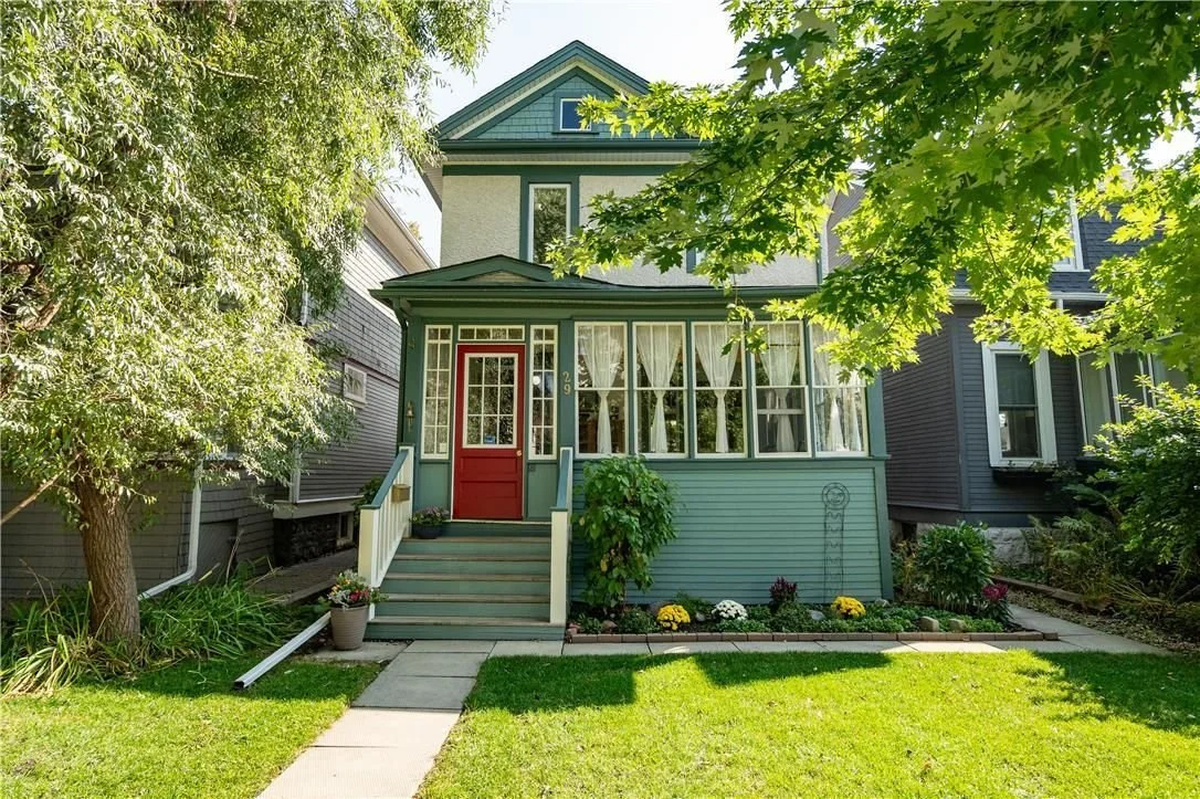 A two-story house painted in light blue with a red front door, white trim, and a porch with decorative curtains, surrounded by greenery and a well-maintained lawn.