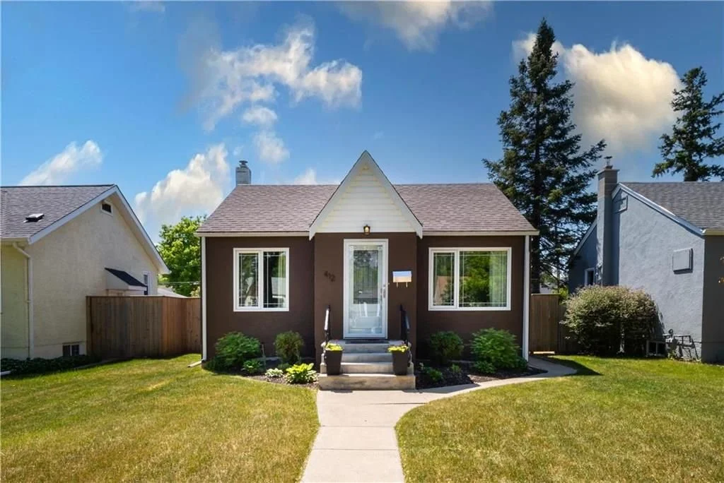 A small brown house with white trim, front porch, and steps, surrounded by a grassy lawn with bushes, in a neighborhood with other similar houses on a sunny day.