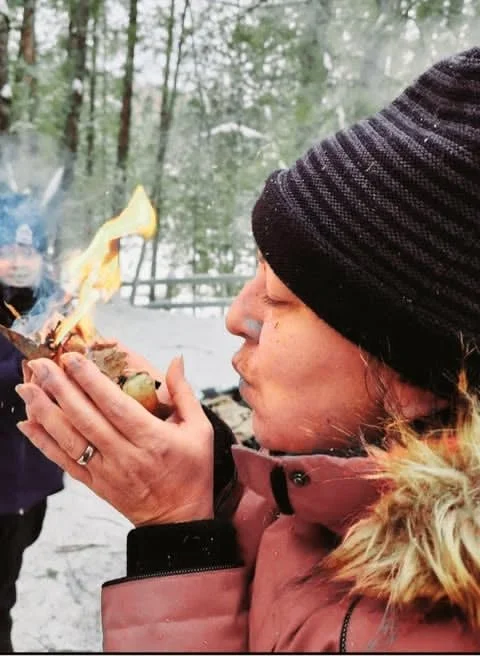 Une femme portant un bonnet noir souffle sur un feu de camp en plein air dans un environnement forestier, avec un autre enfant visible en arrière-plan.