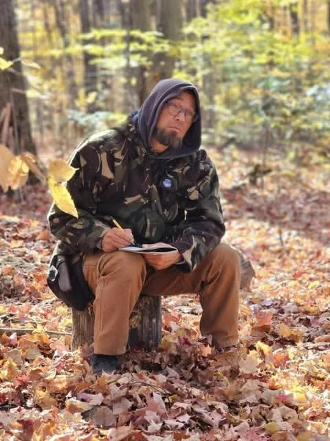 Un homme assis sur un morceau d'arbre dans une forêt en automne, avec des feuilles colorées autour, portant un sweat à capuche gris, un manteau camouflage, et écrit dans un carnet.
