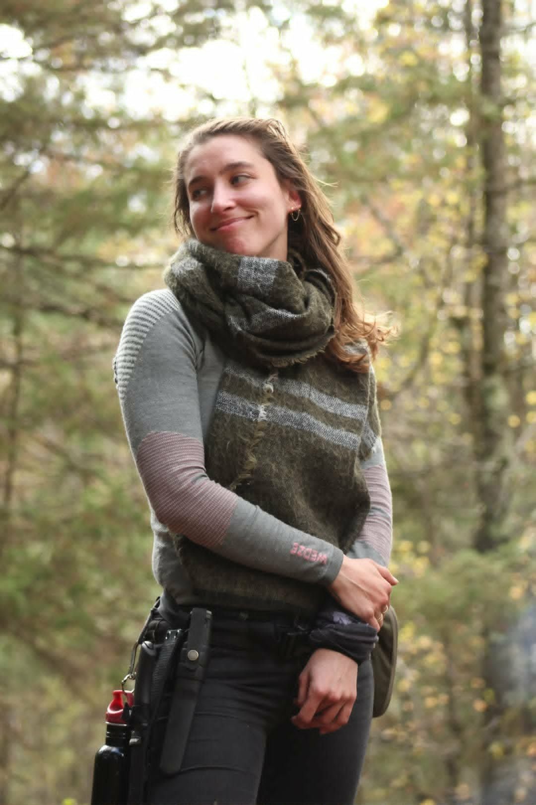 Une femme souriante avec un foulard en laine, portant un pull à manches longues, se tient dans une forêt en automne avec des feuilles colorées en arrière-plan.
