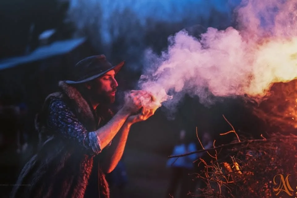 Un homme portant un chapeau souffle de la fumée ou du feu lors d'une soirée en plein air, avec un arrière-plan sombre et quelques personnes en arrière-plan.