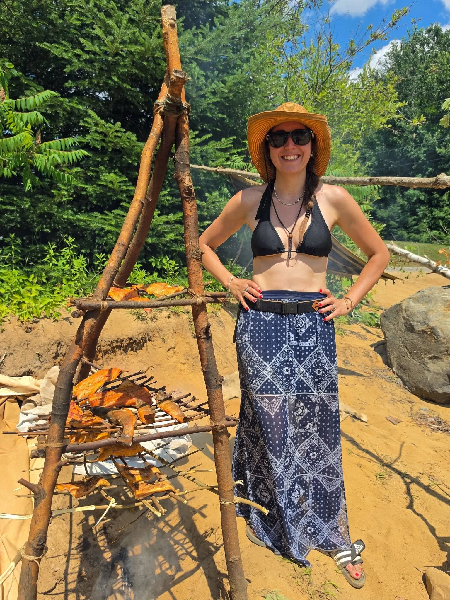 Une femme souriante portant un chapeau en paille, des lunettes de soleil, un haut noir en maillot de bain, et une longue jupe bleue à motifs, se tient à côté d'un feu de bois avec du poisson grillé, dans un environnement naturel avec des arbres et un ciel bleu.