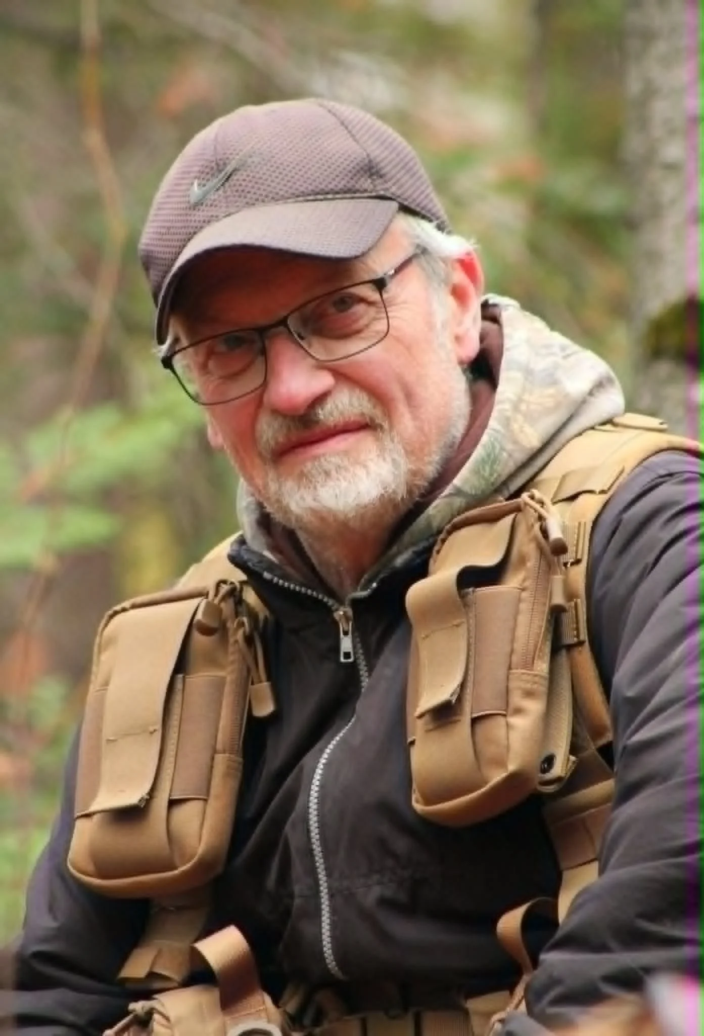 Un homme âgé avec des lunettes, une barbe grise, portant une casquette et un sac à dos beige, dans un environnement naturel forestier.