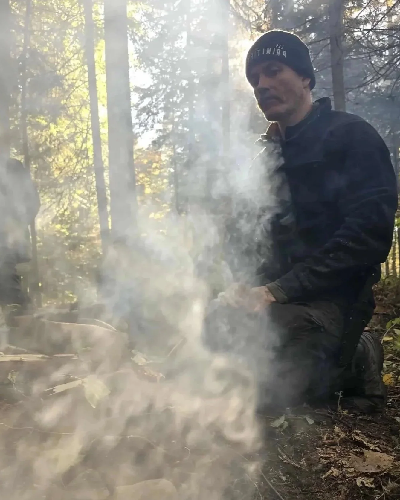Mathieu Hébert en tenue sombre, portant un bonnet noir avec l'inscription en réfléchissant, est agenouillé dans une forêt entourée de fumée de feu de camp.