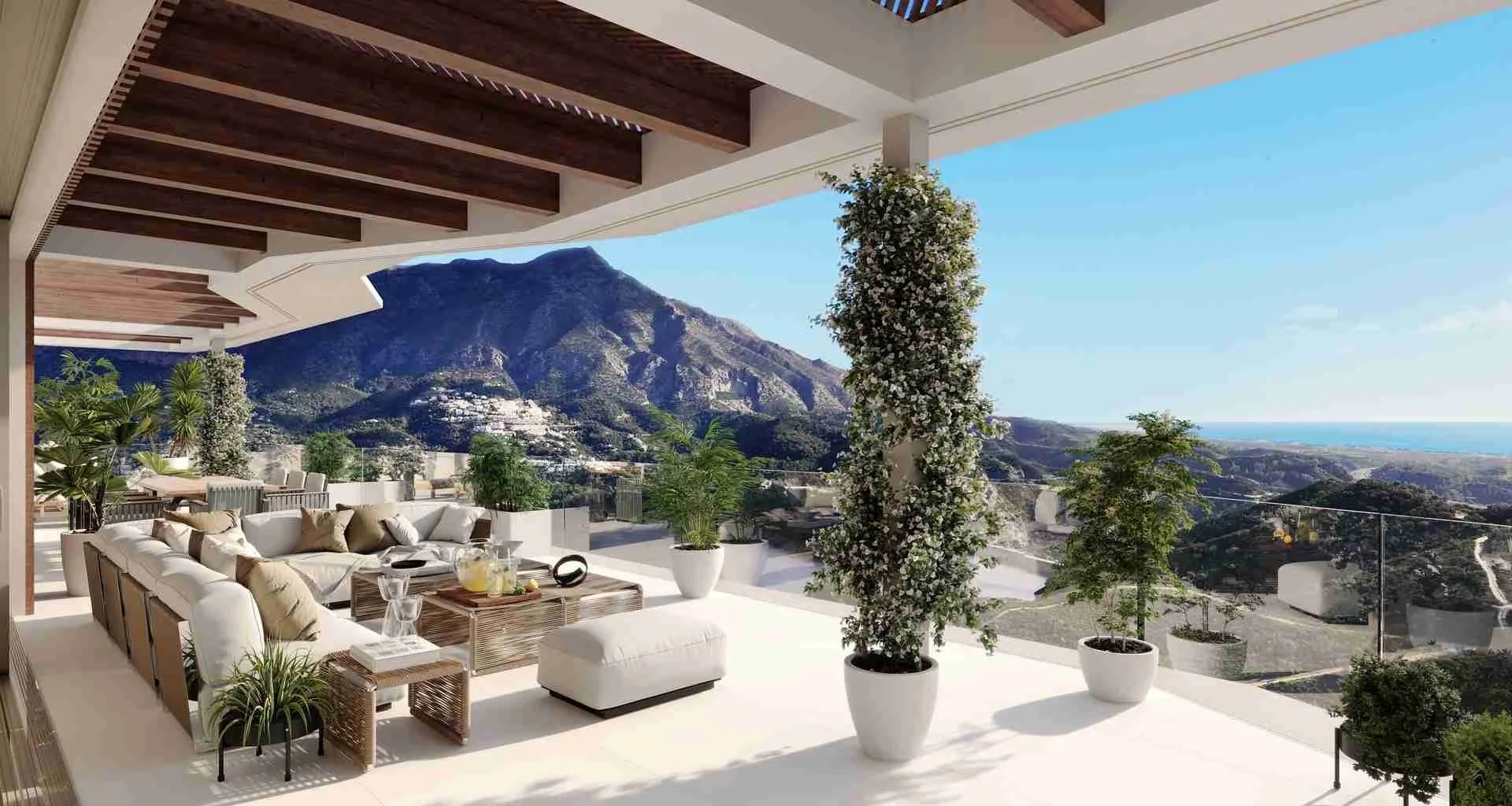 A spacious outdoor terrace with white lounge sofas, a wicker coffee table, and potted plants, overlooking green hills and a mountain with a blue sky in the background.