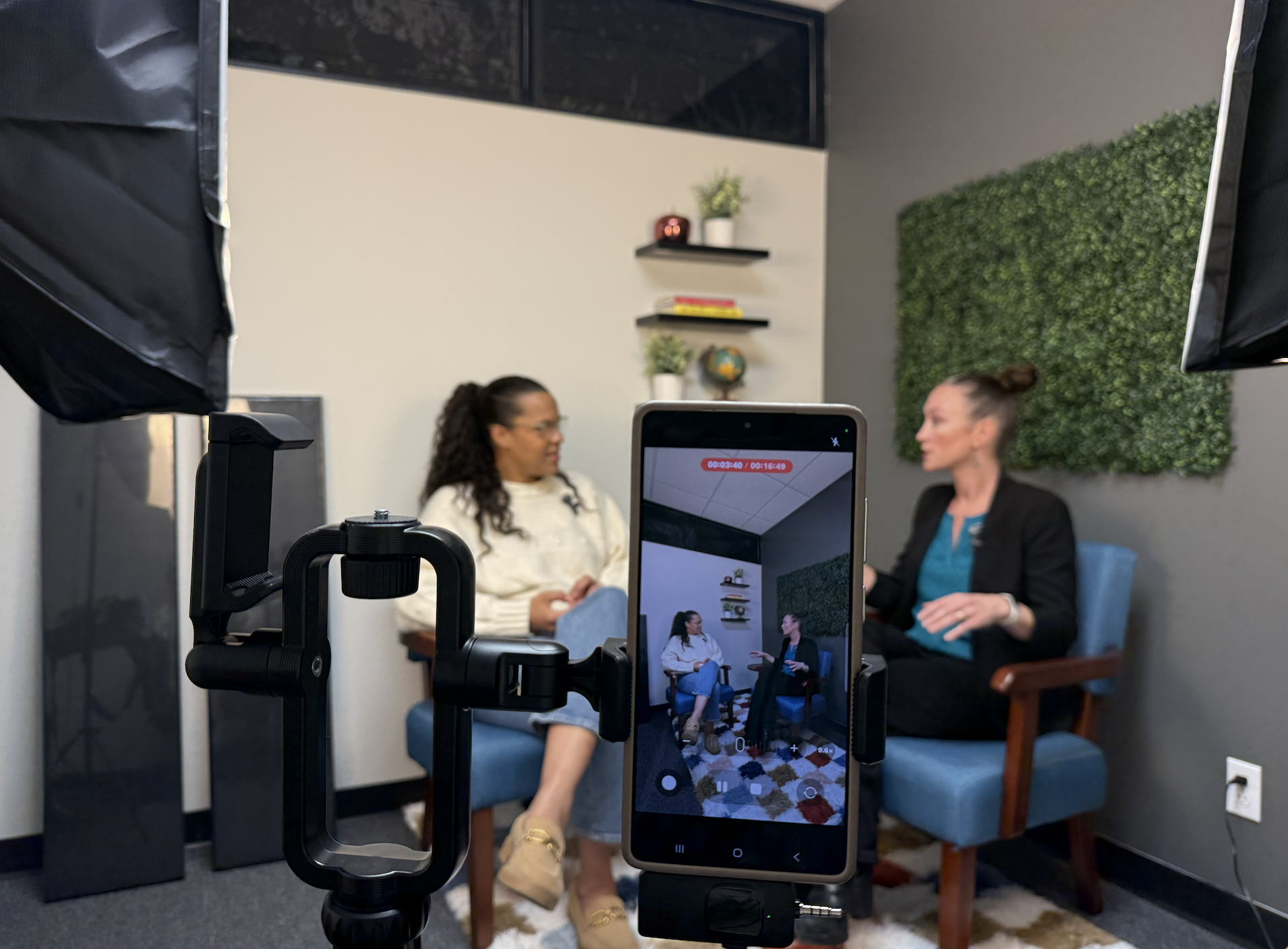 Two women sitting and talking during an interview or discussion in a recording studio. A camera and smartphone are set up to record them, with equipment visible in the foreground. The background features a wall with shelves, a globe, plants, and decorative green wall panels.