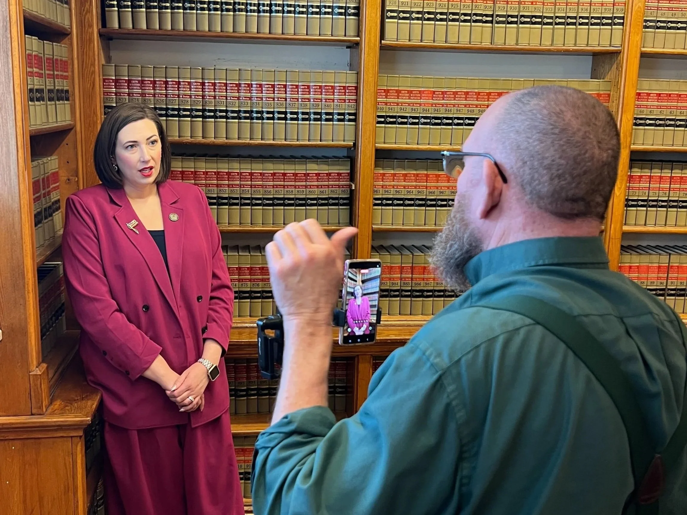 A woman in a pink suit is speaking in front of a camera held by a man with a beard and glasses in a room filled with wood shelves and legal books.