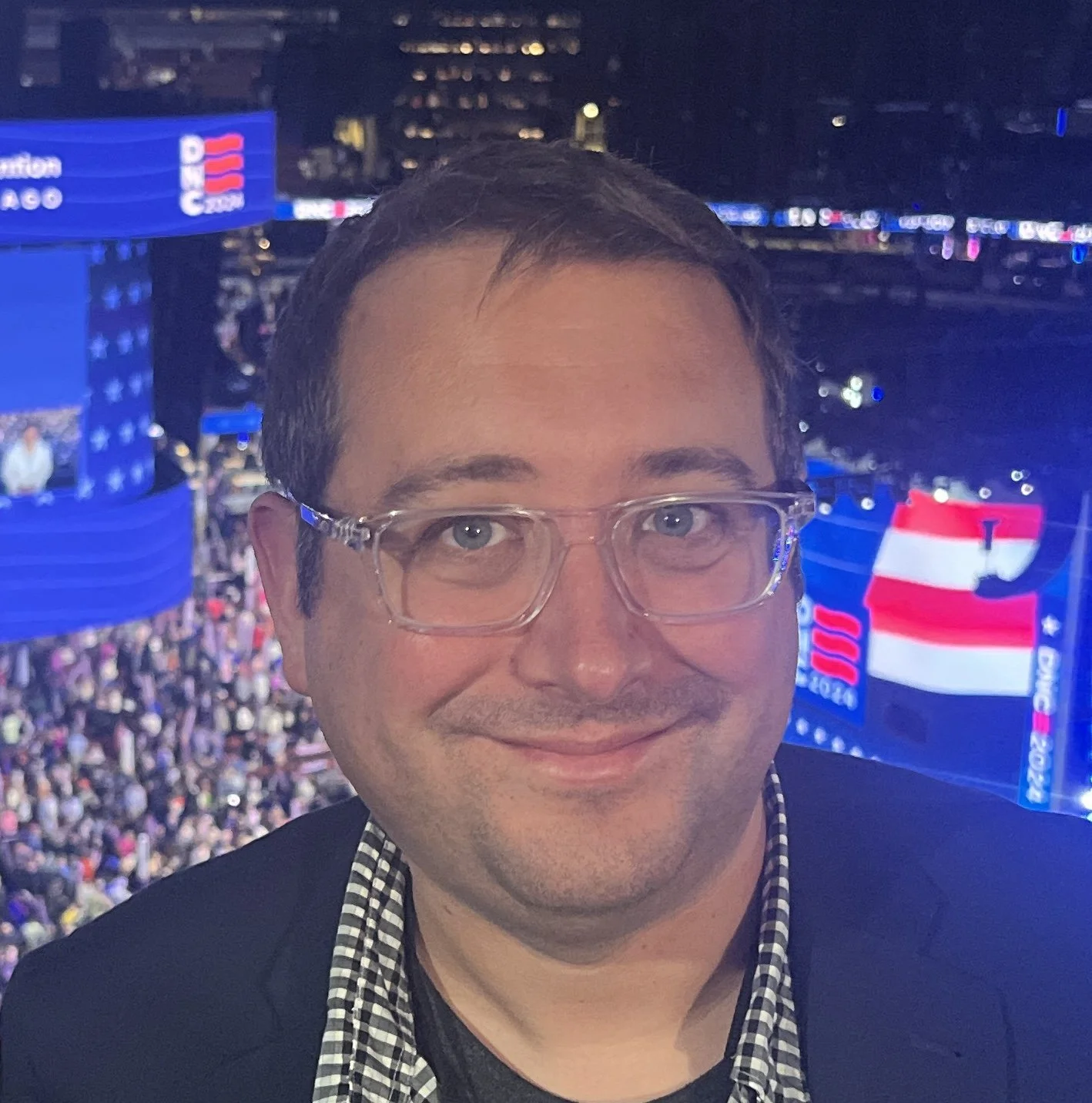 A man smiling in front of a large crowd and multiple screens displaying American flag and event graphics at a political rally or celebration.