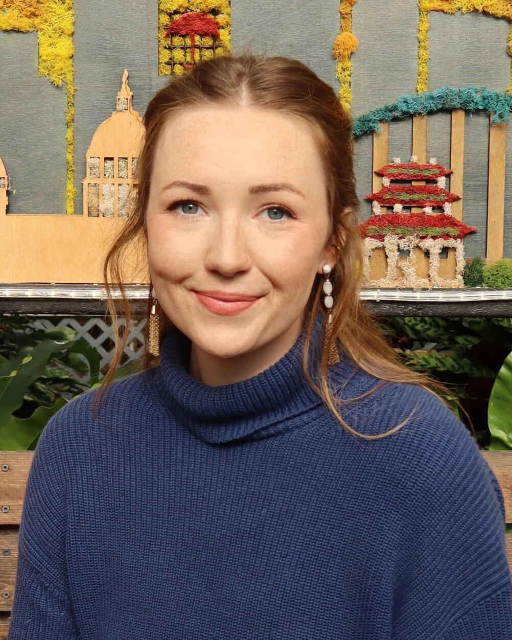 A young woman with red hair, wearing a blue turtleneck sweater and pearl earrings, smiling in front of decorative miniature Japanese buildings and greenery.