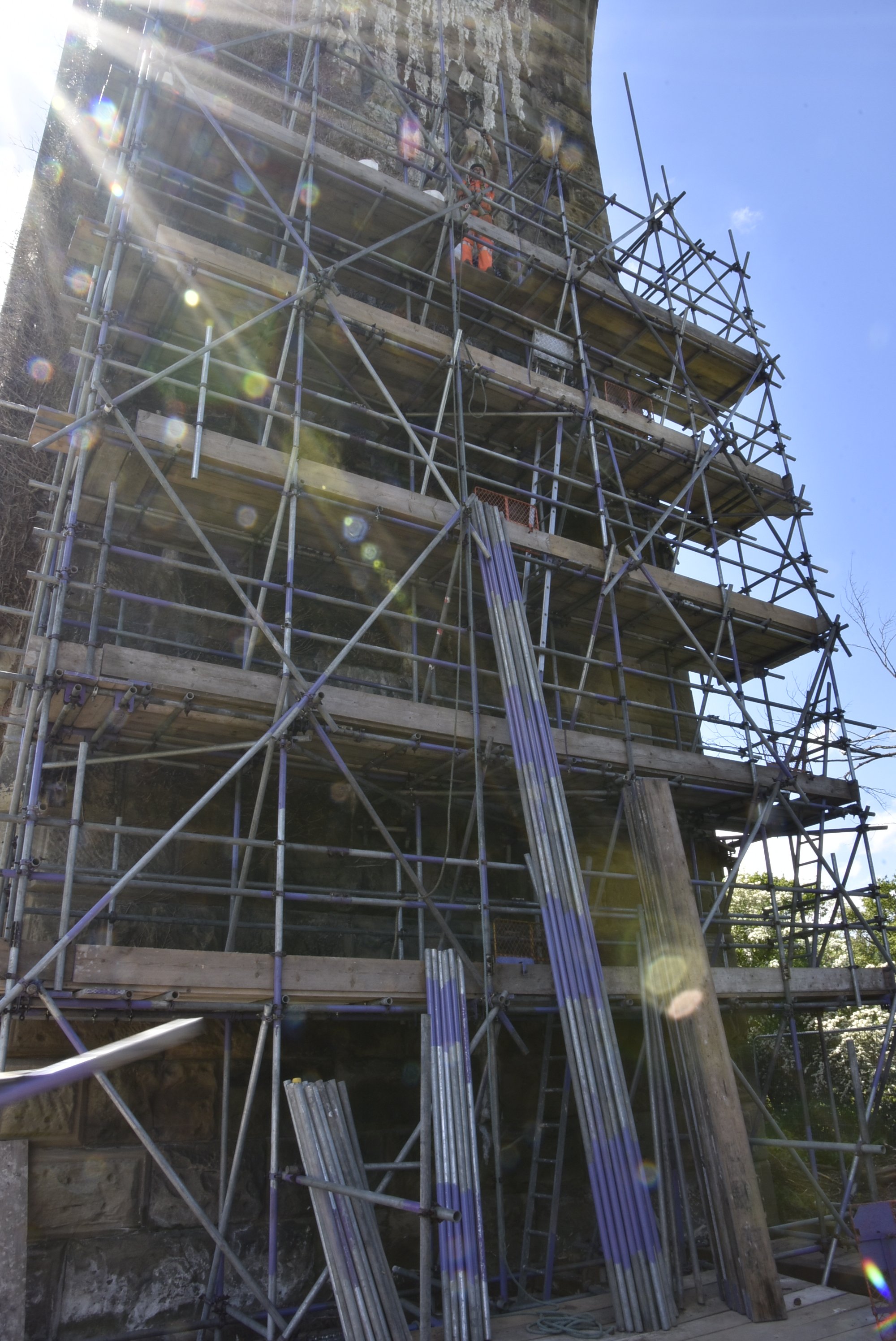 A construction worker in orange safety gear working on scaffolding attached to a large stone structure outdoors under a clear blue sky.