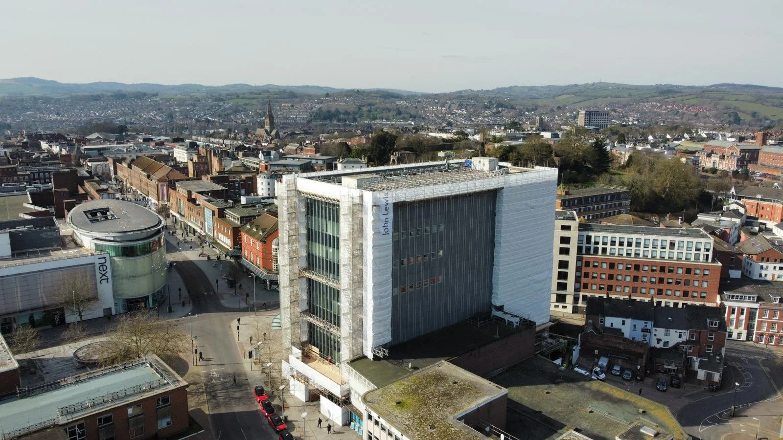 Aerial view of a cityscape featuring modern and historic buildings, a curved shopping mall, with scaffolding