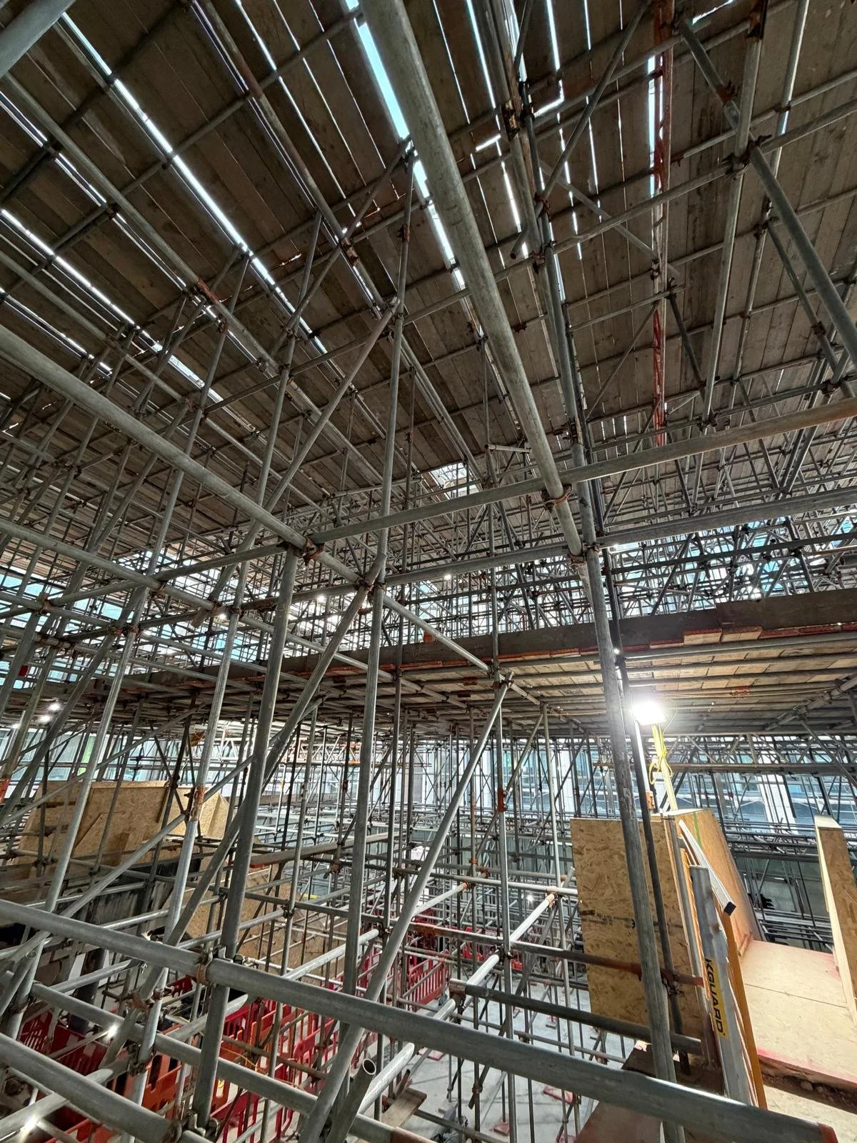 Construction scaffolding inside a building under renovation, with unfinished wooden platforms and metal pipes, and a bright light on a stand.
