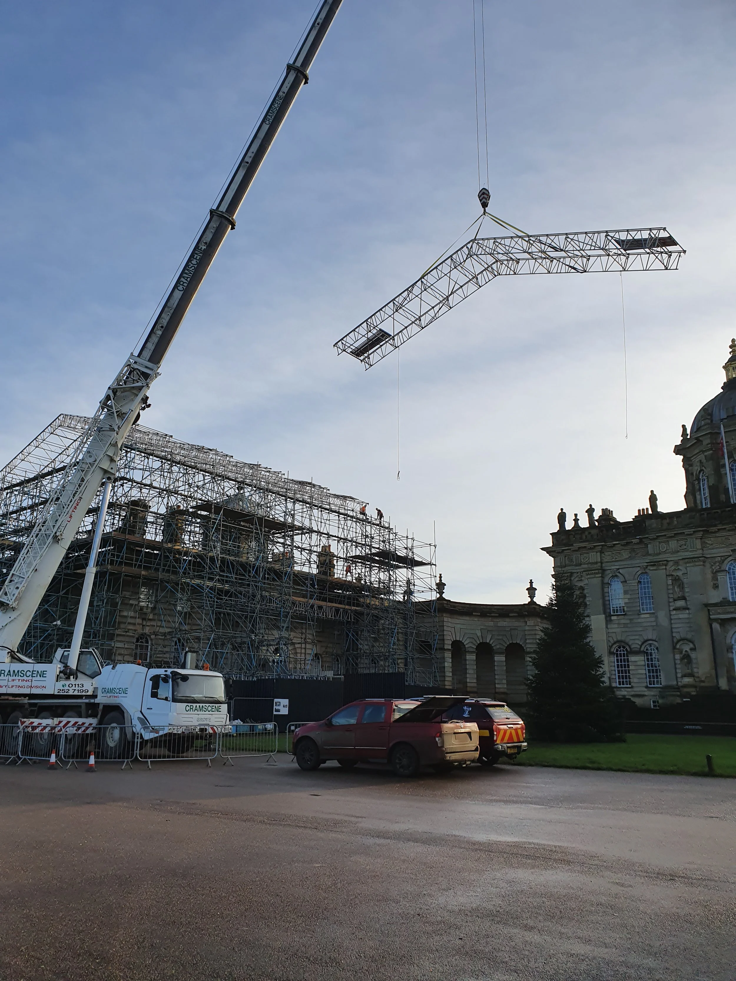 Construction site with cranes installing a large structural framework on a historic building, scaffolding surrounds the building, with parked cars in the foreground, and a clear sky overhead.