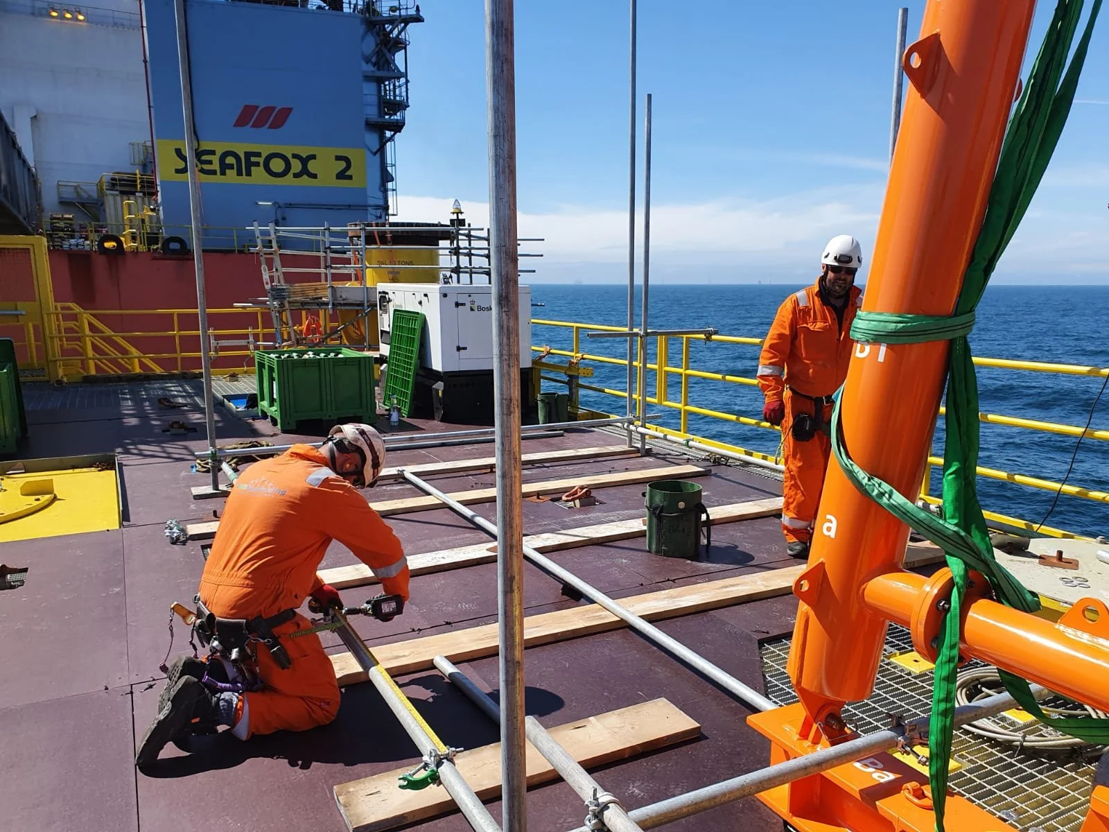 Two workers in orange safety gear and helmets working on a platform on a ship, with the ocean in the background. One worker is kneeling and measuring, the other is standing near a large orange pipe secured with green straps.