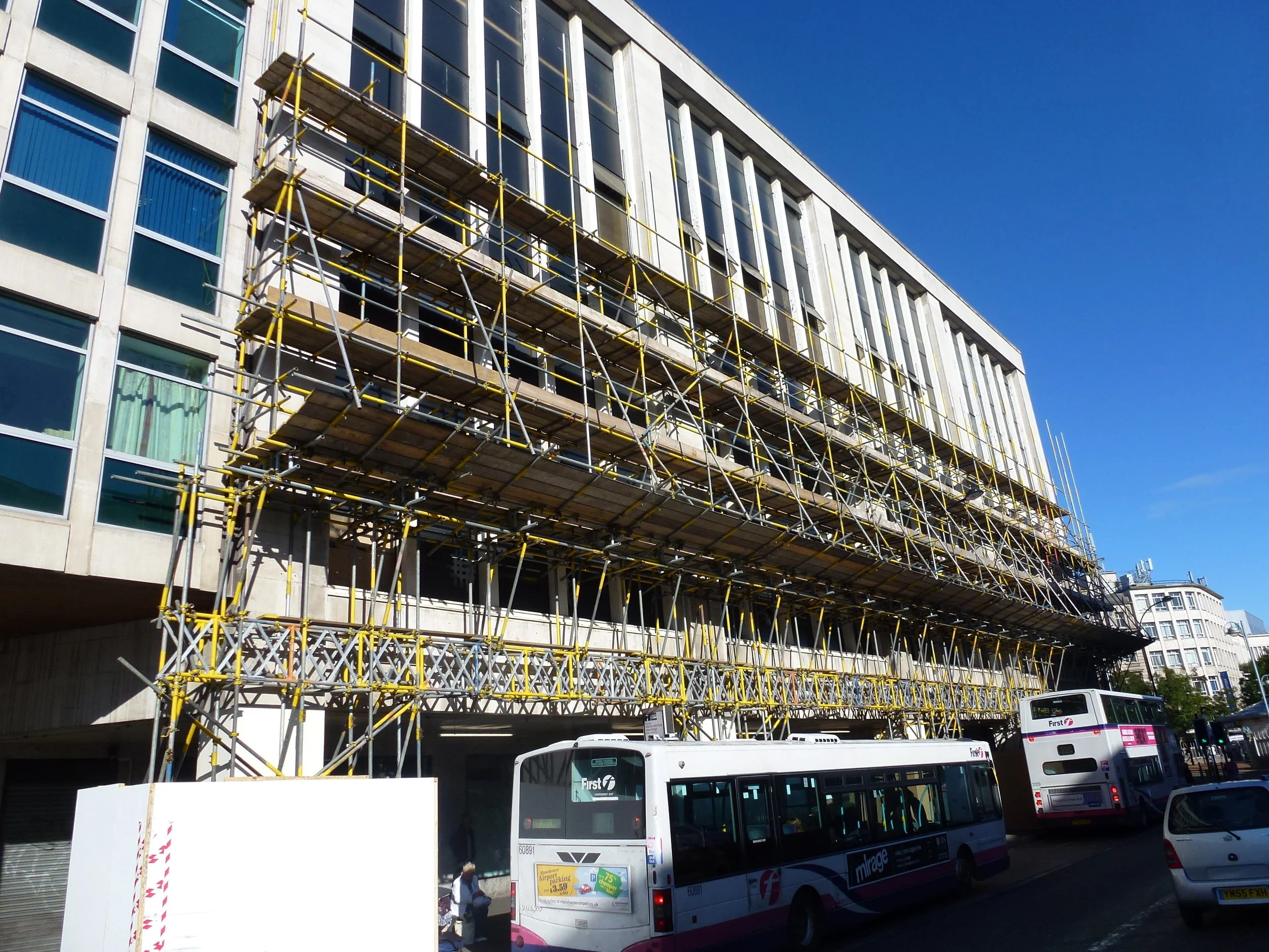 Construction scaffolding on a multi-story building, with two buses and a car parked in front under a clear blue sky.