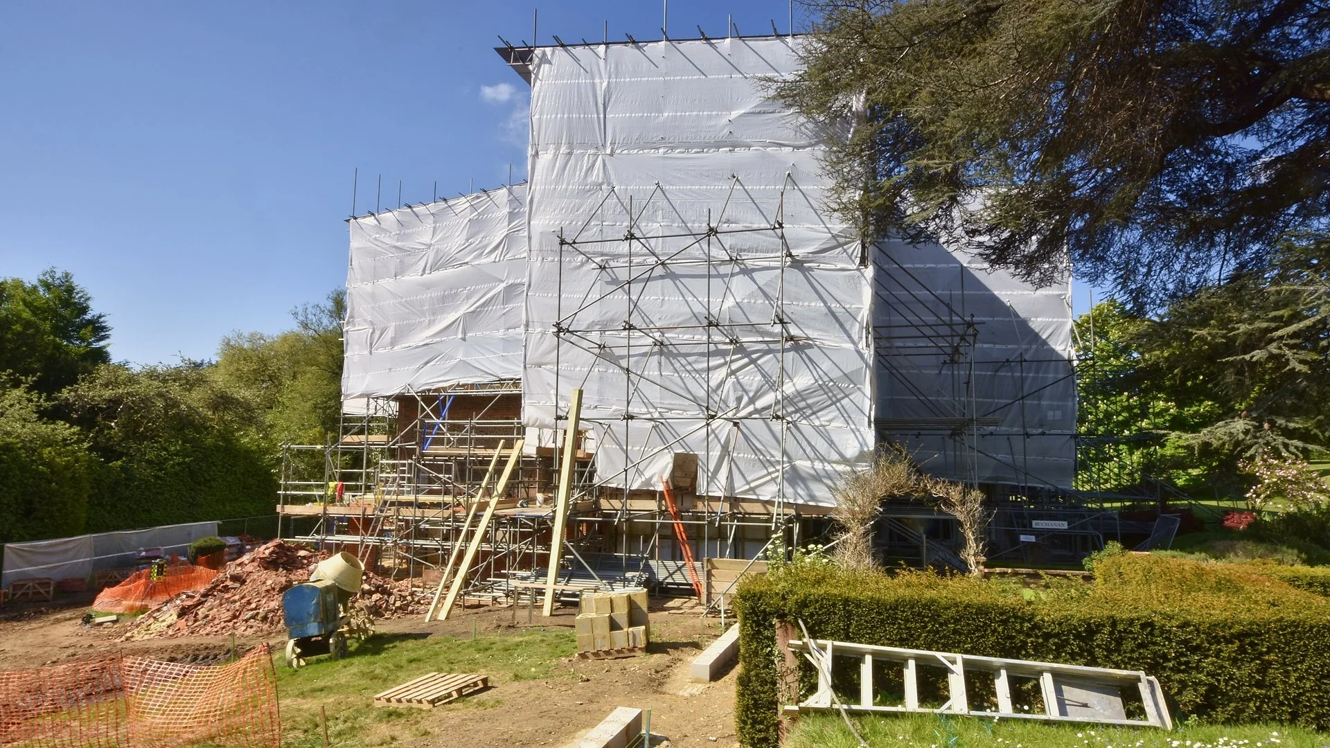 Construction site with scaffolding and white protective coverings on a building under renovation, surrounded by trees and green landscape under a blue sky.