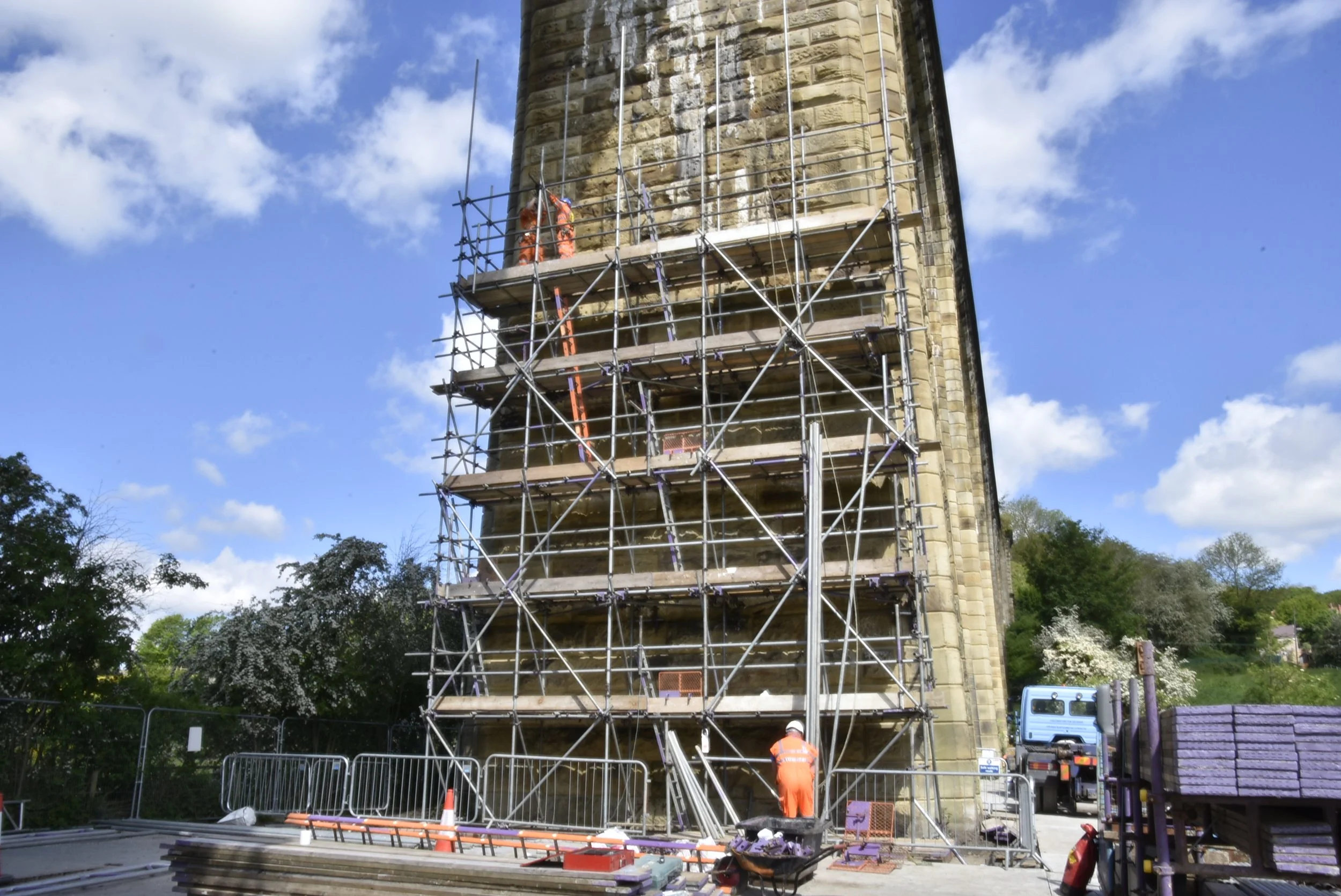 Workers in orange safety uniforms repairing a tall stone tower surrounded by scaffolding, with a clear blue sky and some clouds in the background.