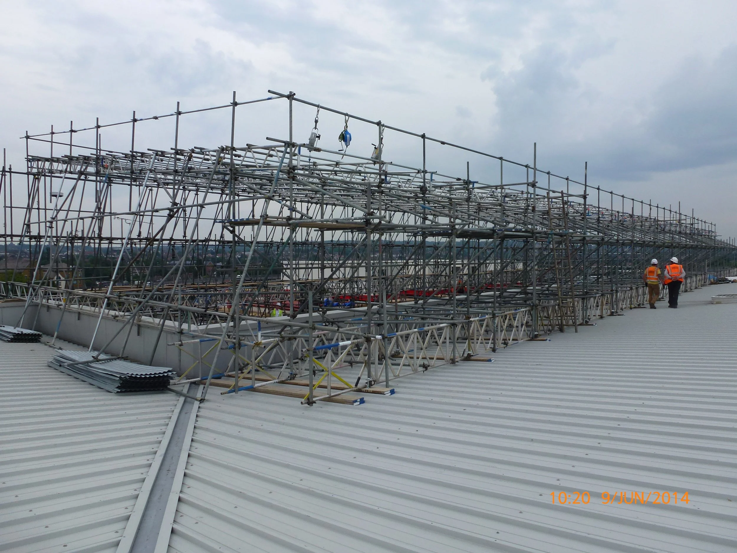 Construction workers in safety vests and helmets walking on a metal roof with metal scaffolding and framework installed on top in an overcast sky.
