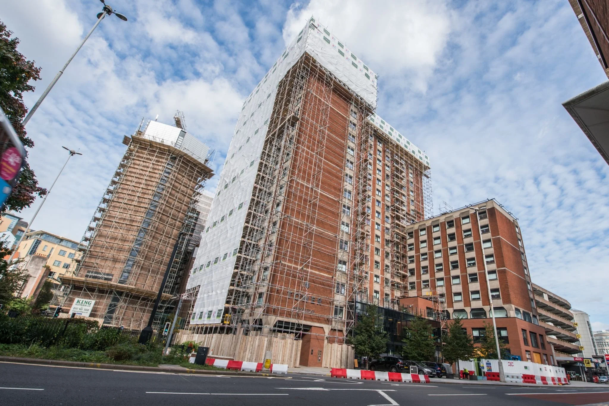 Construction site with high-rise buildings under renovation, scaffolding wrapped around the buildings, and a clear partly cloudy sky.