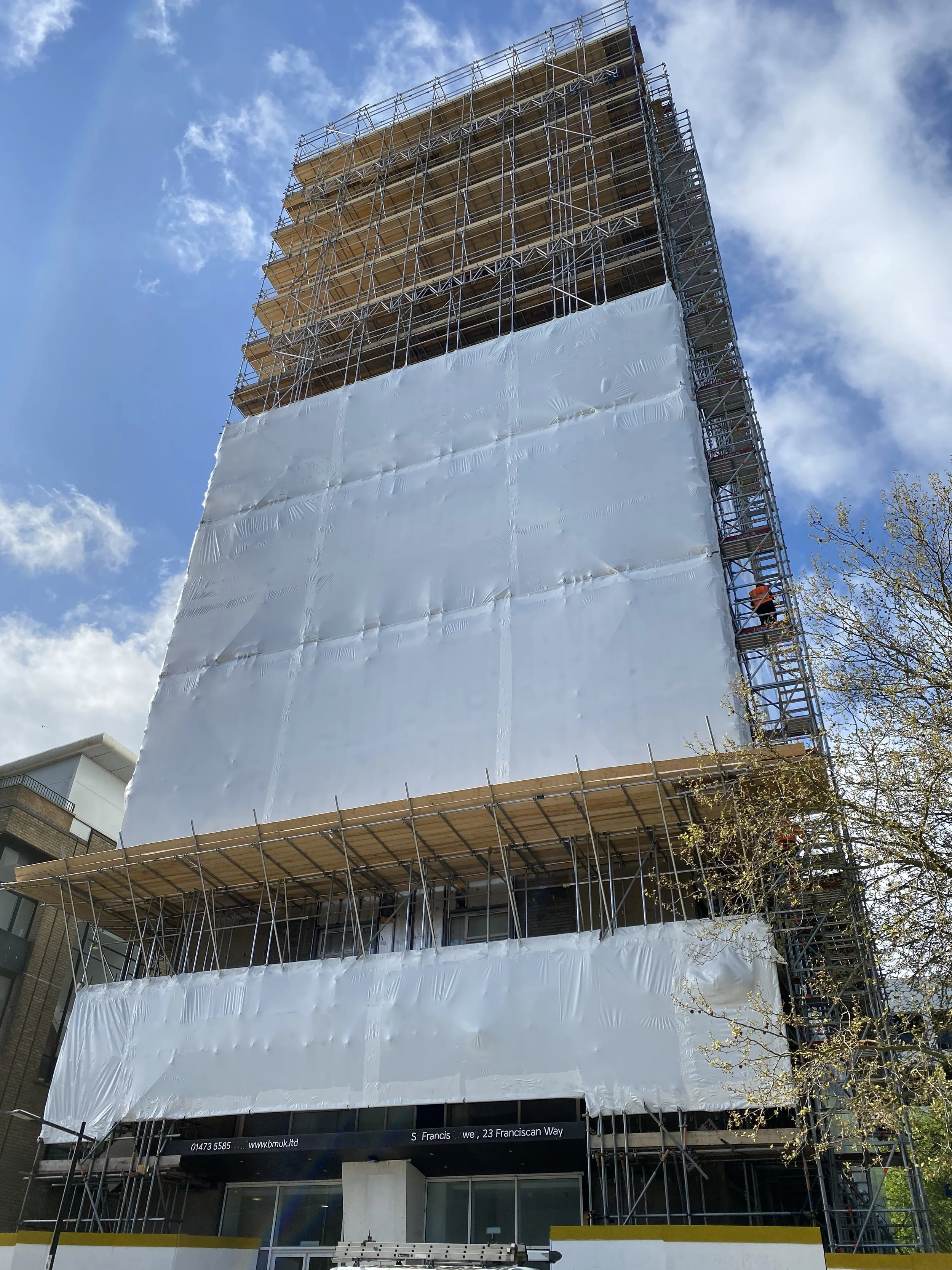 A tall building under construction covered with scaffolding and white protective sheeting, with clear blue sky and clouds overhead.