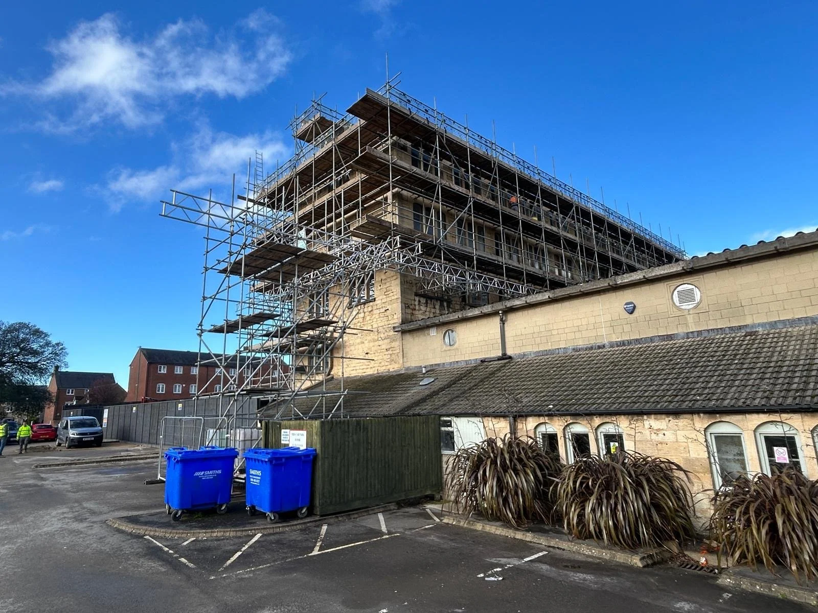 A multi-story building under renovation with scaffolding, surrounded by a parking lot that has a few people, vehicles, and blue trash bins.