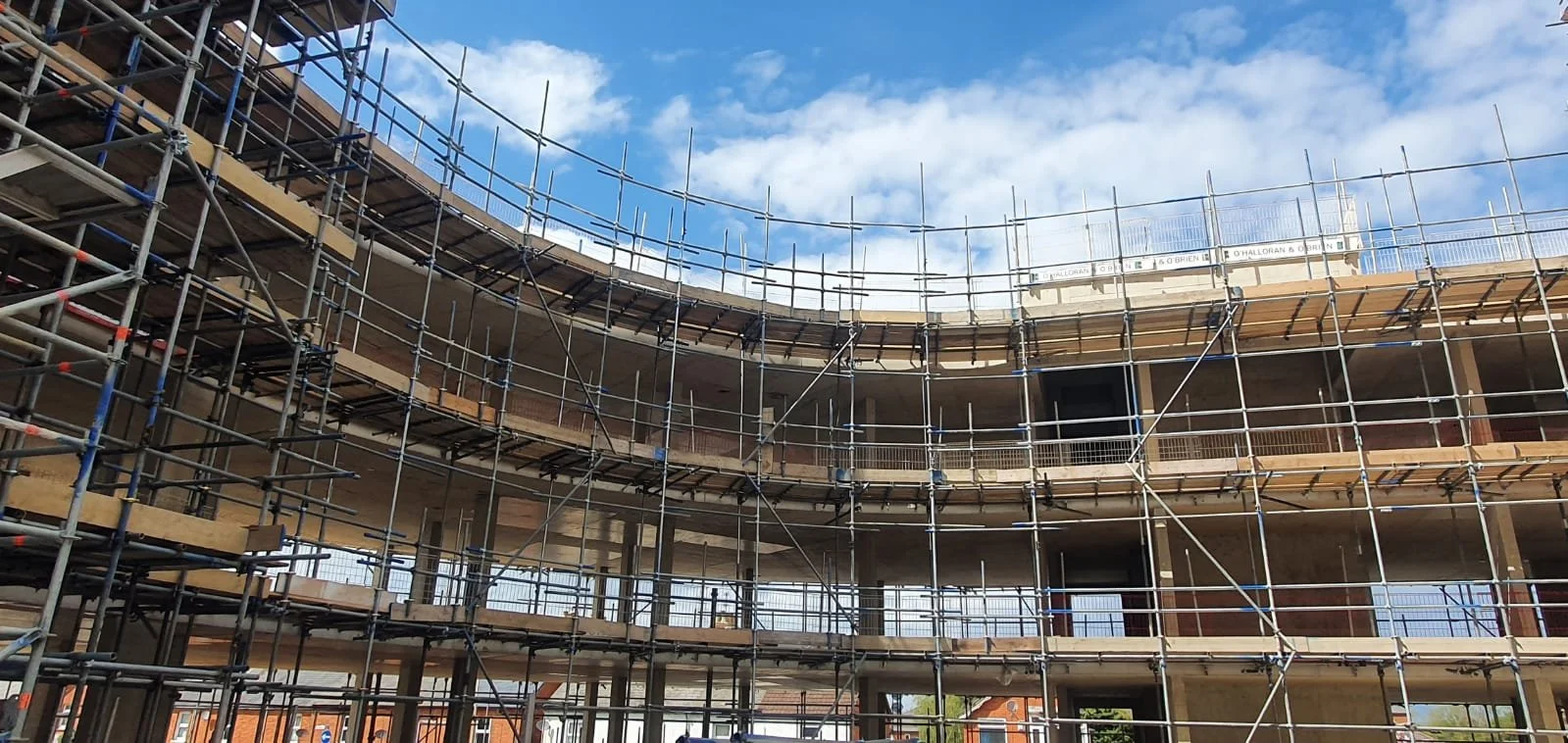 Construction scaffolding surrounds a multi-story building under construction with an open framework and a cloudy blue sky overhead.
