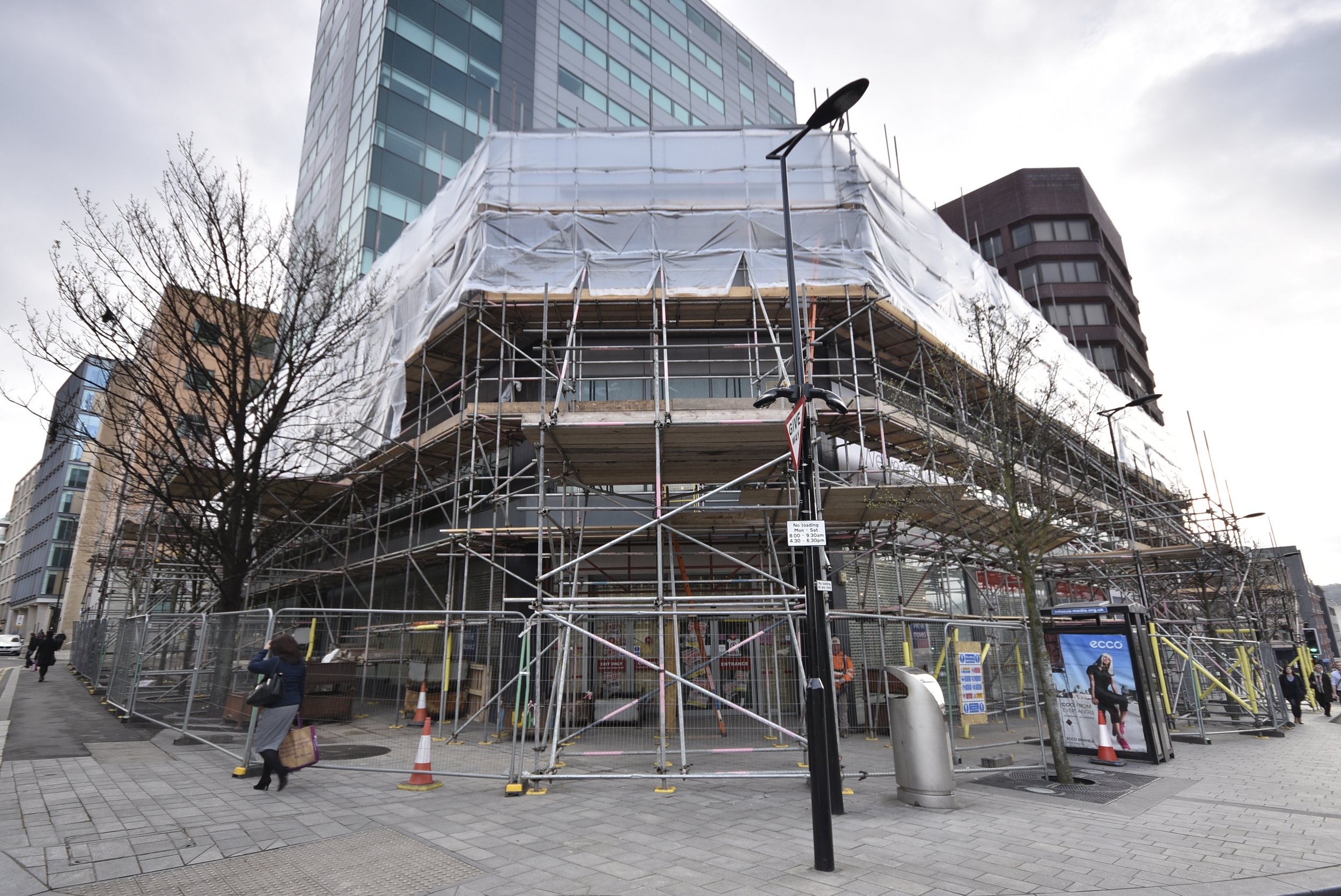 A building under construction surrounded by metal scaffolding and white protective sheeting, located on a city street with pedestrians and leafless trees.
