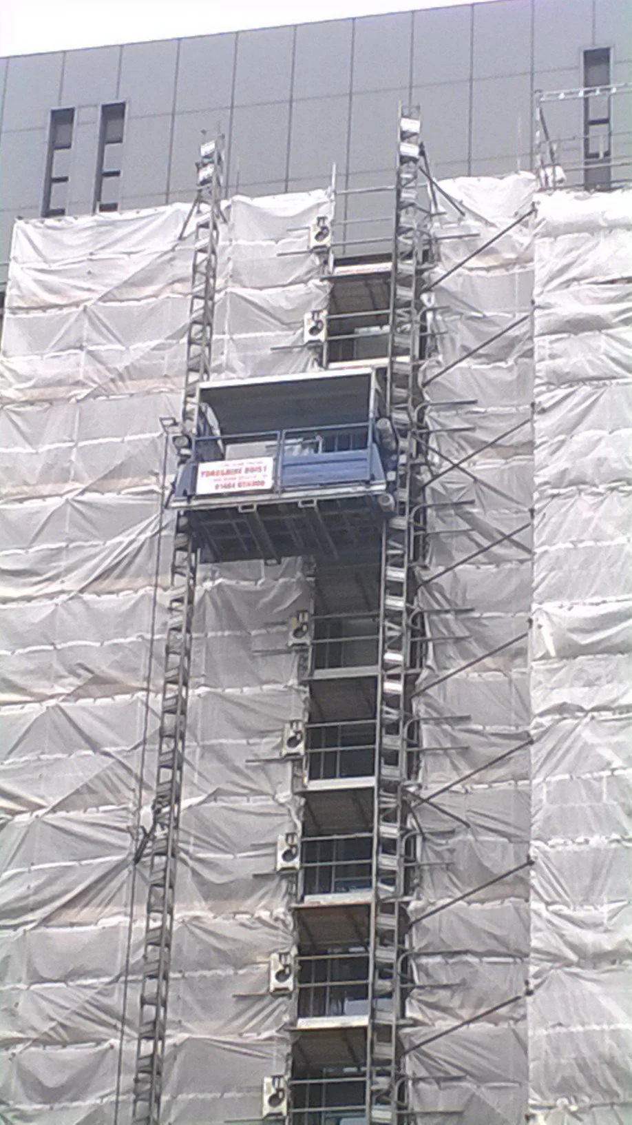 Construction scaffolding and wrapping on a building under renovation with a suspended platform and a safety sign.