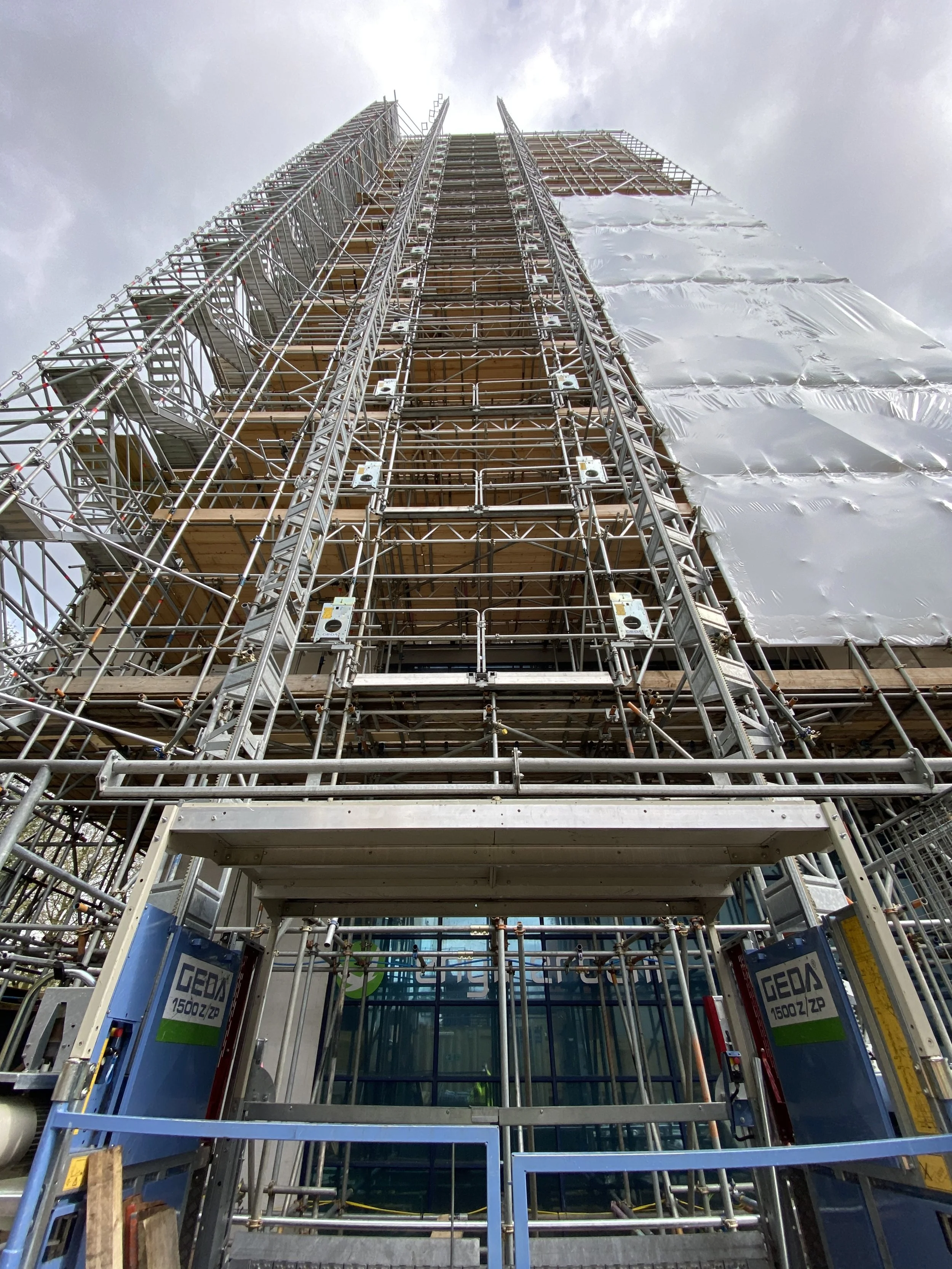 A tall building under construction covered with scaffolding and white protective sheeting, viewed from the ground looking upward.