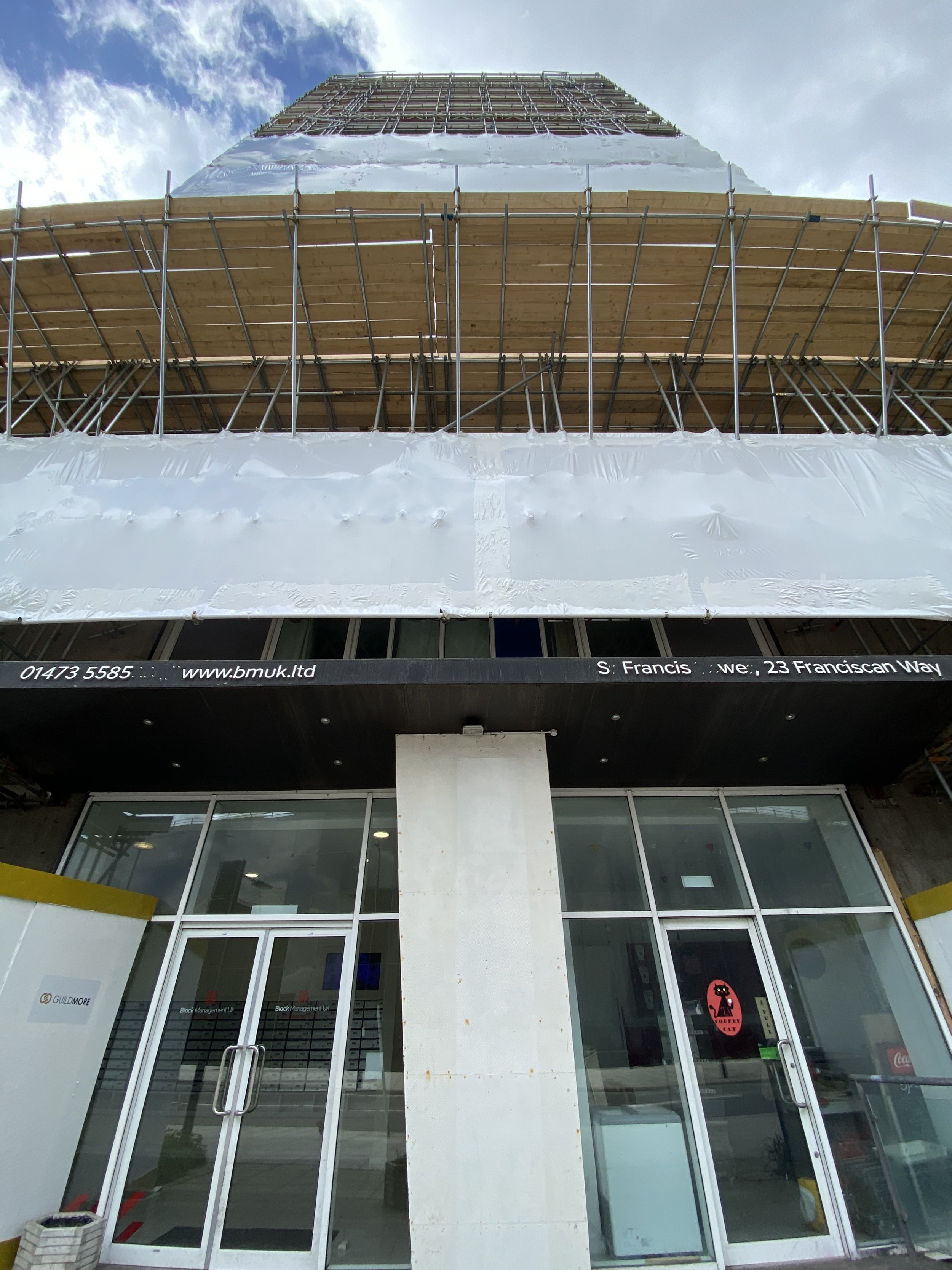 View of a building under construction with scaffolding on the middle and upper levels, and a glass entrance on the ground floor.