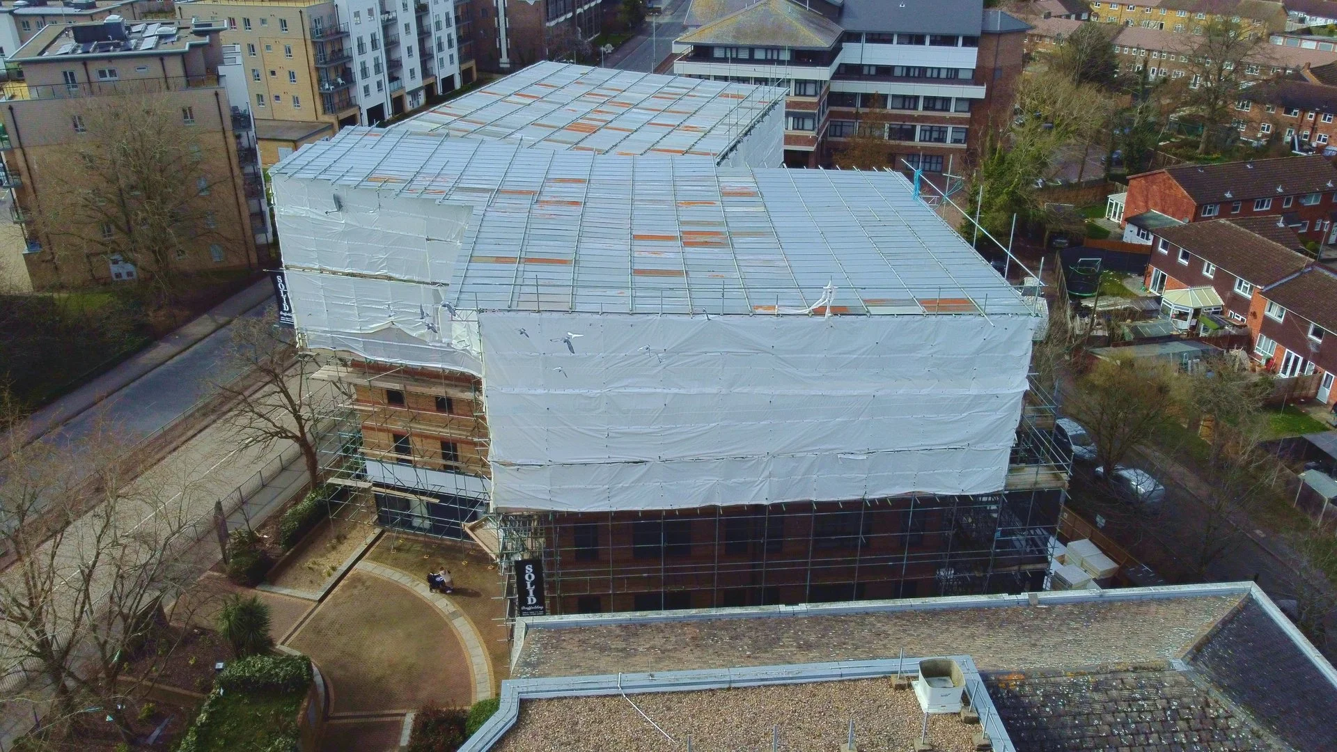 Aerial view of a multi-story building under construction with white scaffolding and cover, surrounded by residential buildings and trees.
