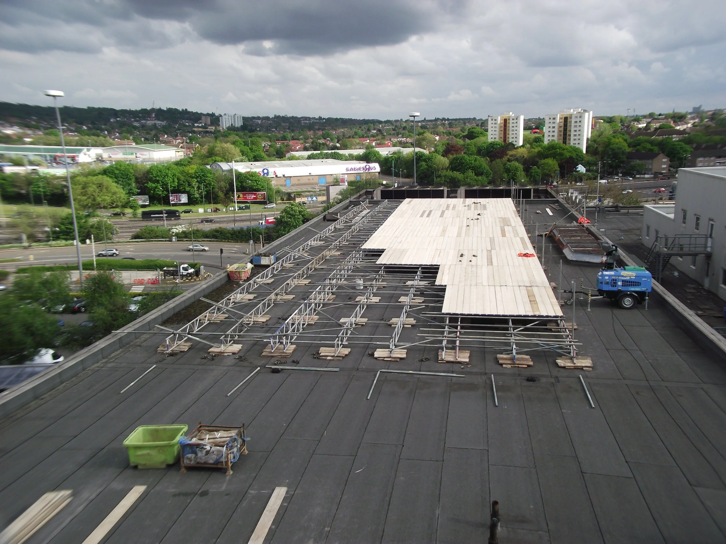 View from a rooftop showing a flat roof under construction with metal framework, wooden panels, construction tools, and equipment, with a cityscape of trees, buildings, and parking lots in the background under an overcast sky.