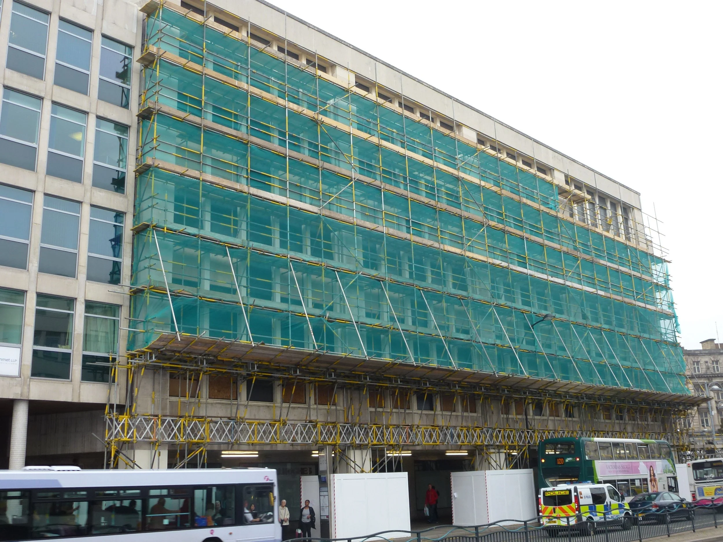 A building under construction with scaffolding and green safety netting, city street scene with buses, cars, and pedestrians.