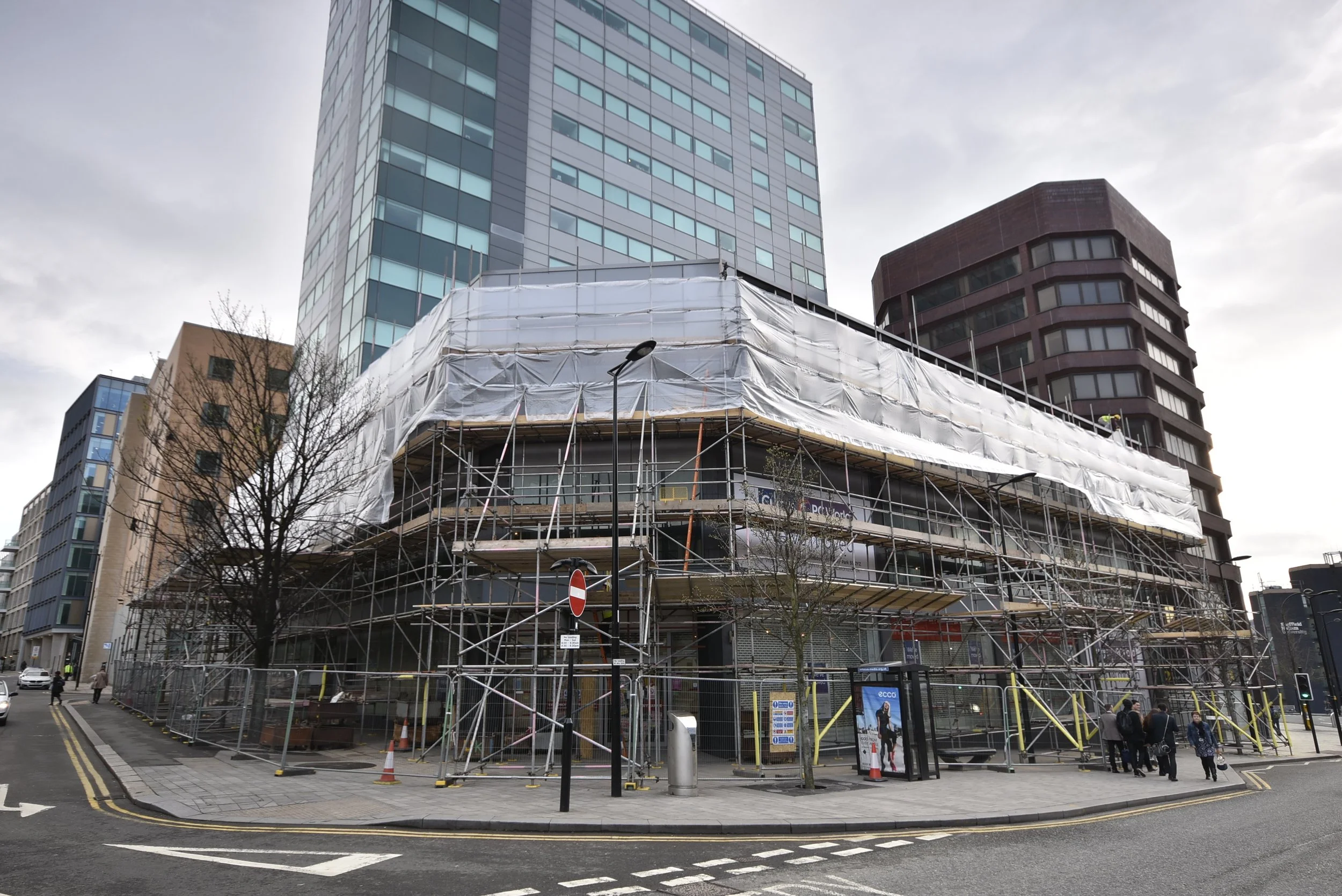 Construction site with scaffolding surrounding a building on a city street, with pedestrians and a bus stop nearby.