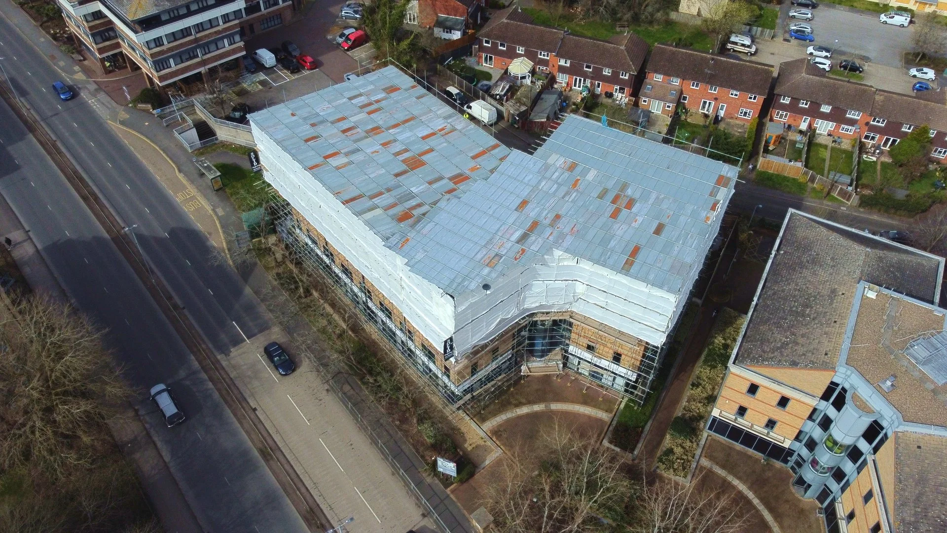 An aerial view of a building under construction, with scaffolding around it and a metal roof in progress, located near residential houses and a street with parked cars.