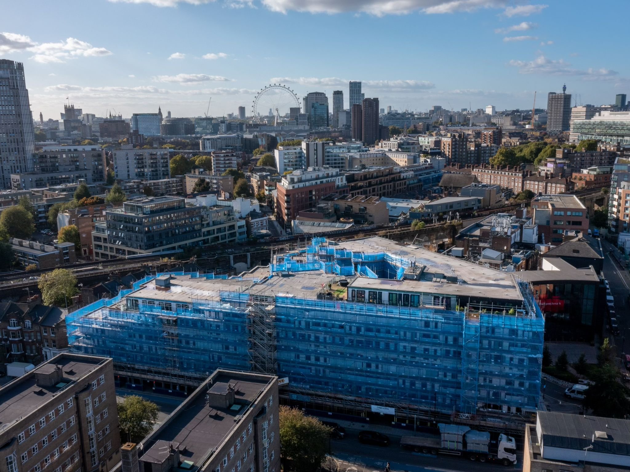 A cityscape view of London featuring a construction site with blue scaffolding in the foreground, and the London Eye, skyscrapers, and historic buildings in the background on a partly cloudy day.