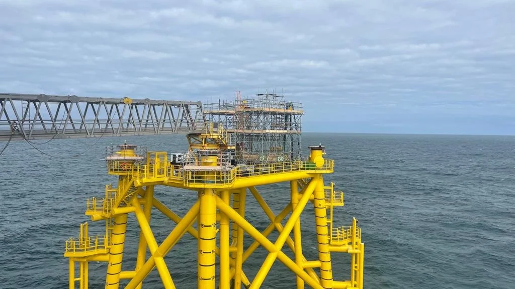 Offshore oil drilling platform with yellow structural components in the ocean under a cloudy sky.