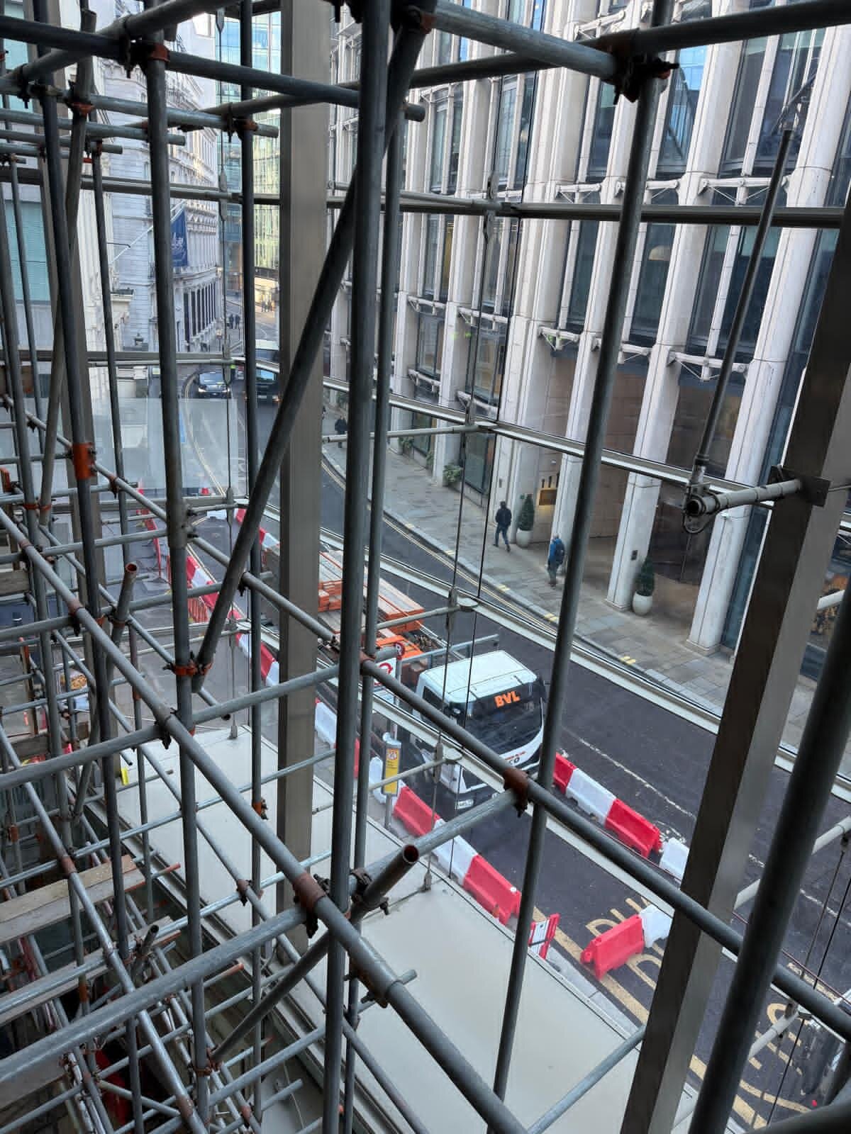 View from a building under construction showing metal scaffolding, a city street with construction barriers, a bus, pedestrians, and tall buildings.