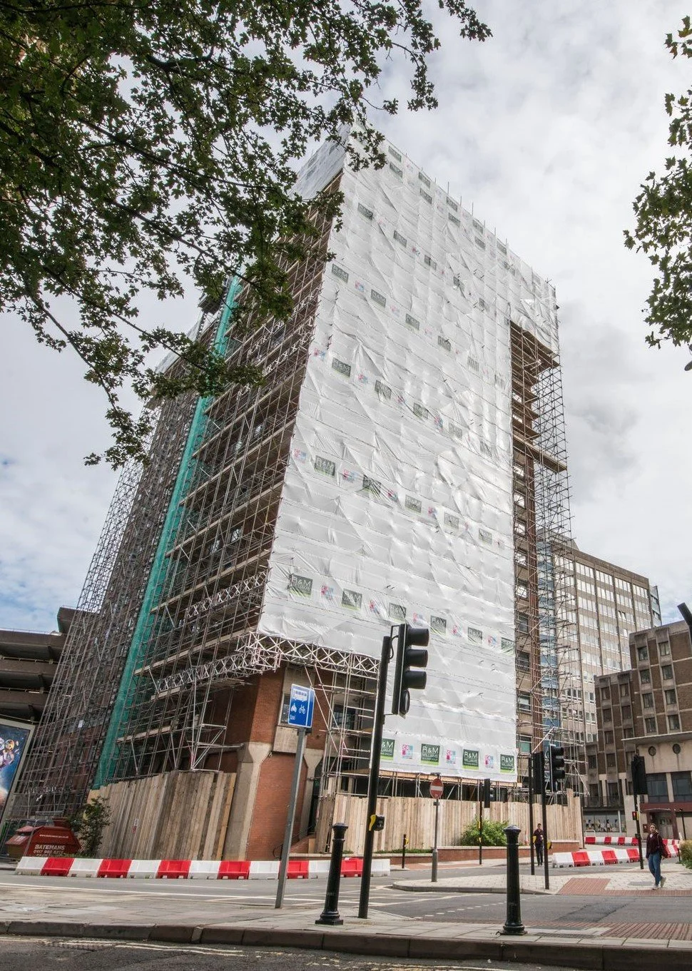 A tall building under construction wrapped in white protective sheeting, with scaffolding visible on the sides. There are trees in the foreground and street signs, including a bicycle lane sign, and pedestrians near the street.