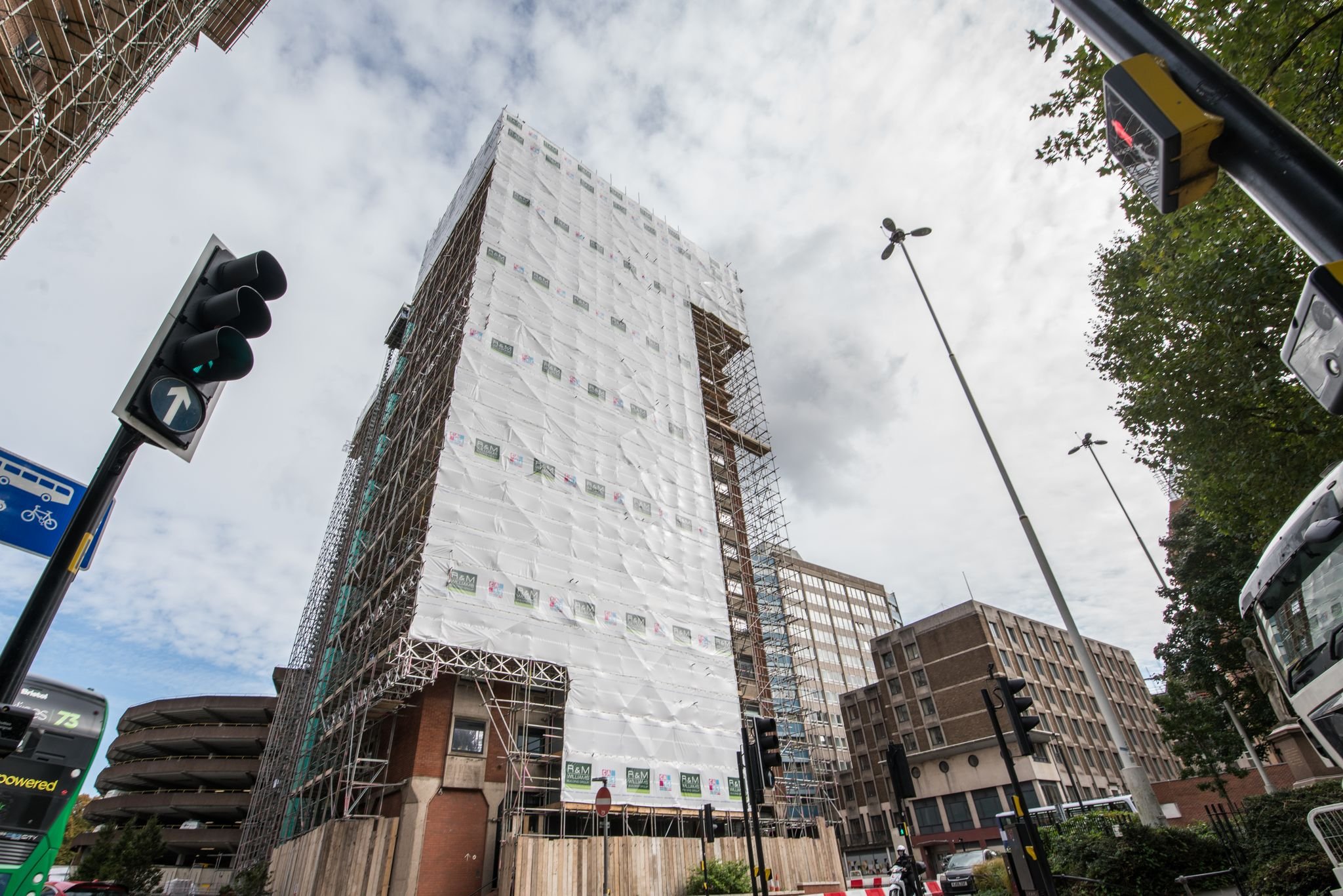 Under construction high-rise building surrounded by scaffolding and white construction wrap, with traffic lights, street lamps, a bus and city buildings in the background, under cloudy sky.