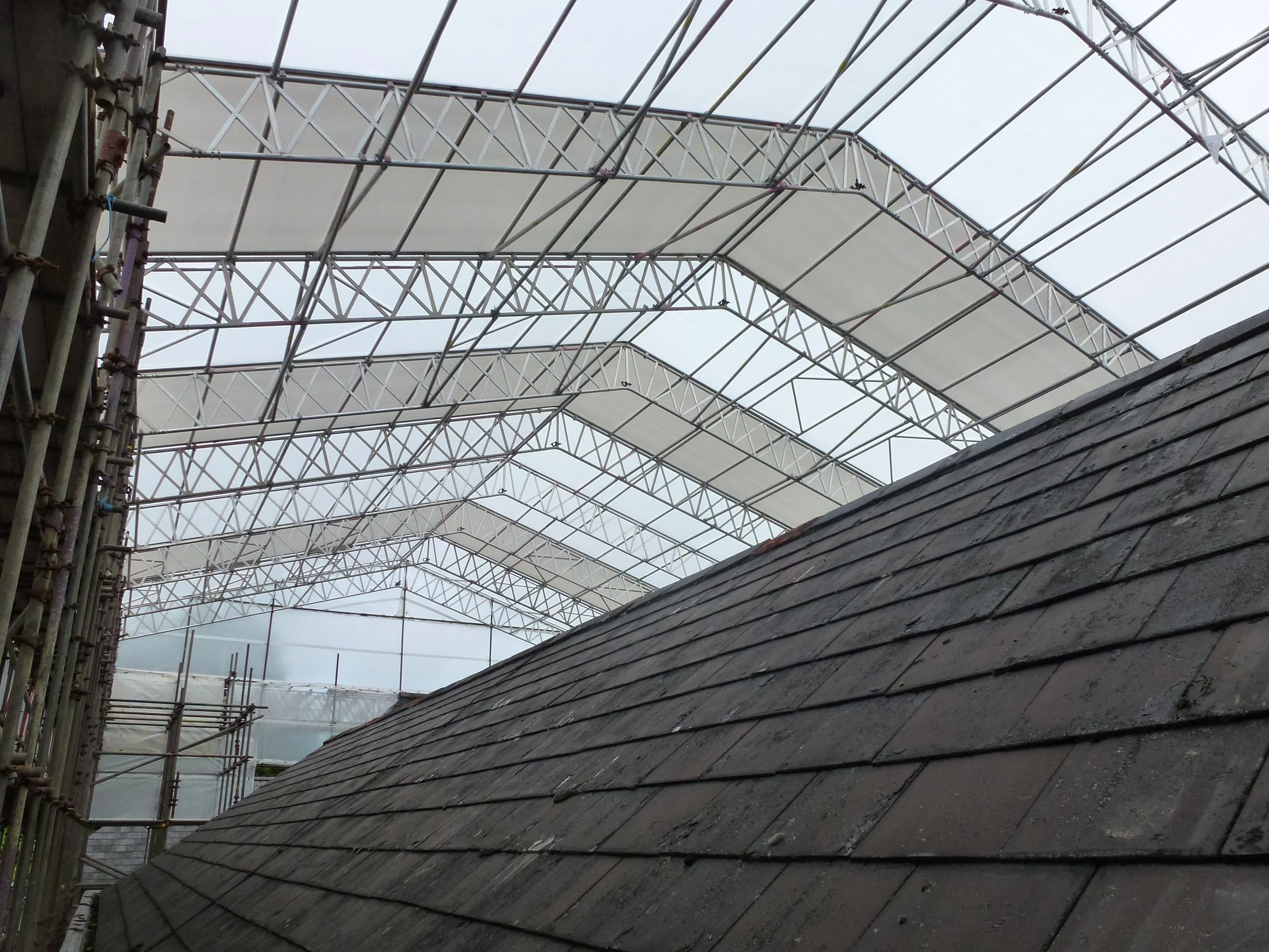 Construction site with a dark gray shingled roof and a transparent canopy supported by metal scaffolding.