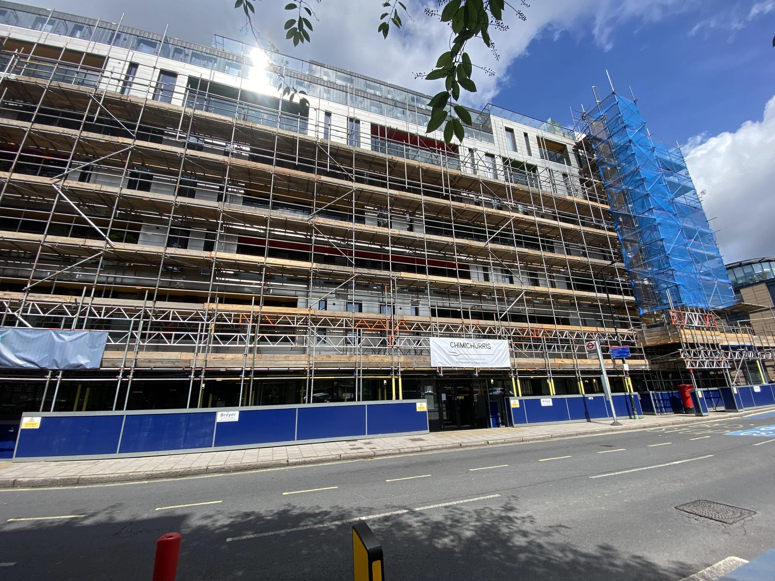 A multi-story building under construction with scaffolding, blue safety netting on one side, and a blue construction fence at the ground level. The building has glass windows and white exterior walls, and the photo shows a partly cloudy sky with sunl